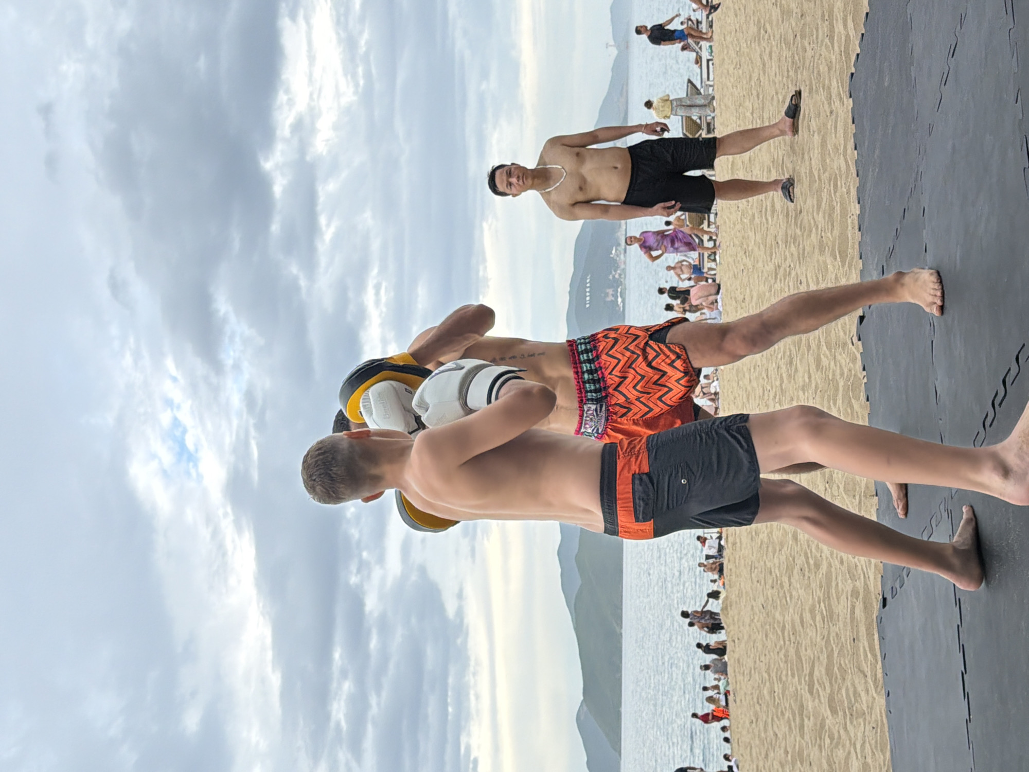 Two individuals are engaged in what appears to be a Muay Thai training session on a black mat placed on a sandy beach. The person in the foreground, a young Caucasian male with blonde hair, wears dark gray shorts with an orange trim and is holding focus mitts, actively participating in the training. The second individual, a Caucasian male with darker hair, is wearing patterned orange shorts and is striking the focus mitts. In the background, several other people are present on the beach, some are seated on loungers, and others are standing near the water. The sea stretches out to the horizon, with a mountainous coastline visible in the distance. The sky is overcast with gray clouds. The scene is likely located on a public beach in Nha Trang, Vietnam, a popular tourist destination known for its coastline and water activities.