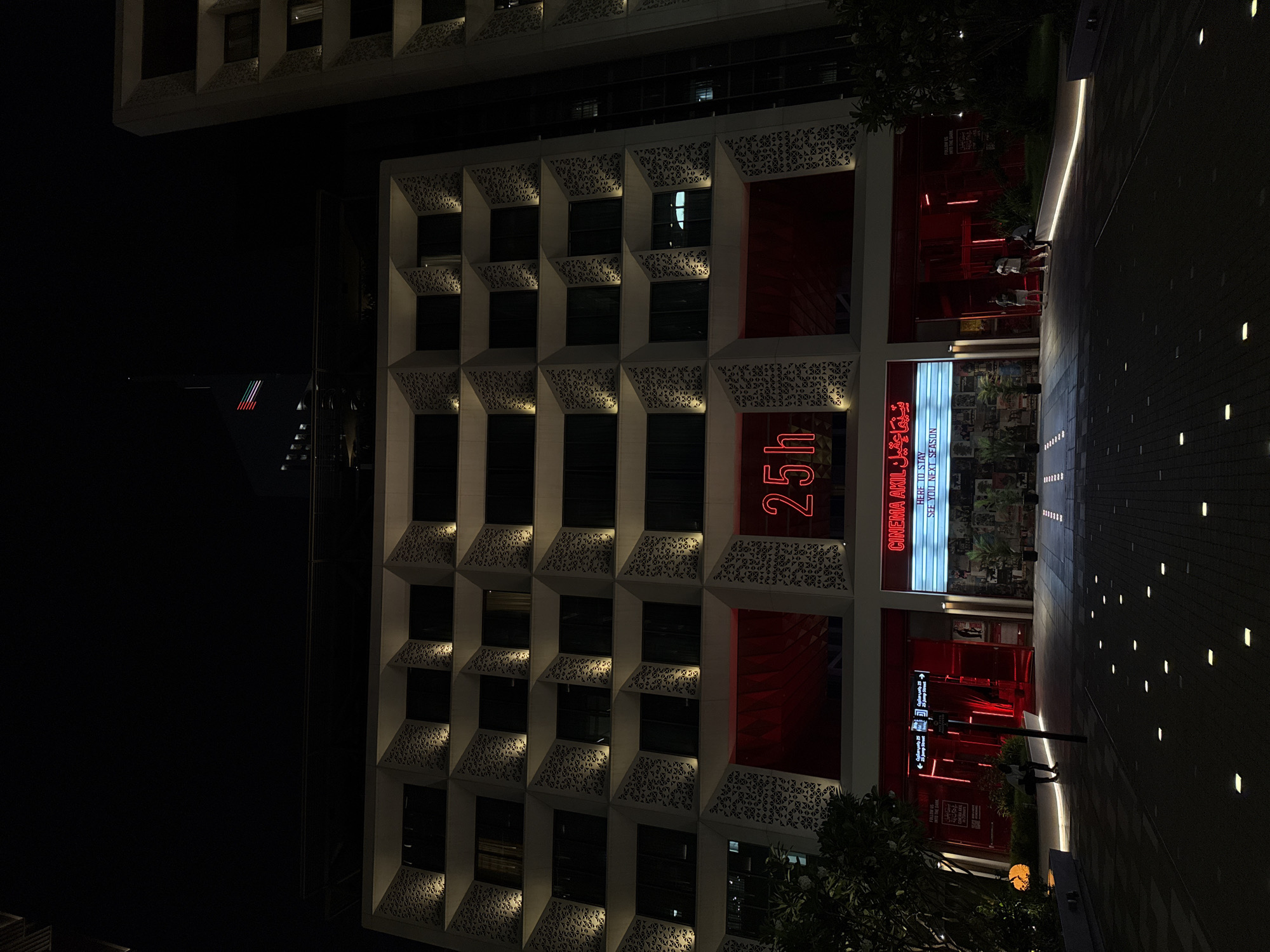 The image shows a building at night in Dubai, United Arab Emirates, with a modern facade featuring geometric patterns around its window frames. A sign reading "25h" is lit up on the building's exterior, next to a cinema named "Cinema Akil." The cinema's signage includes text in Arabic and English, indicating it is "HERE TO STAY" and inviting patrons to "SEE YOU NEXT SEASON." People are present on the sidewalk, and there is a walkway with lights embedded in the pavement.