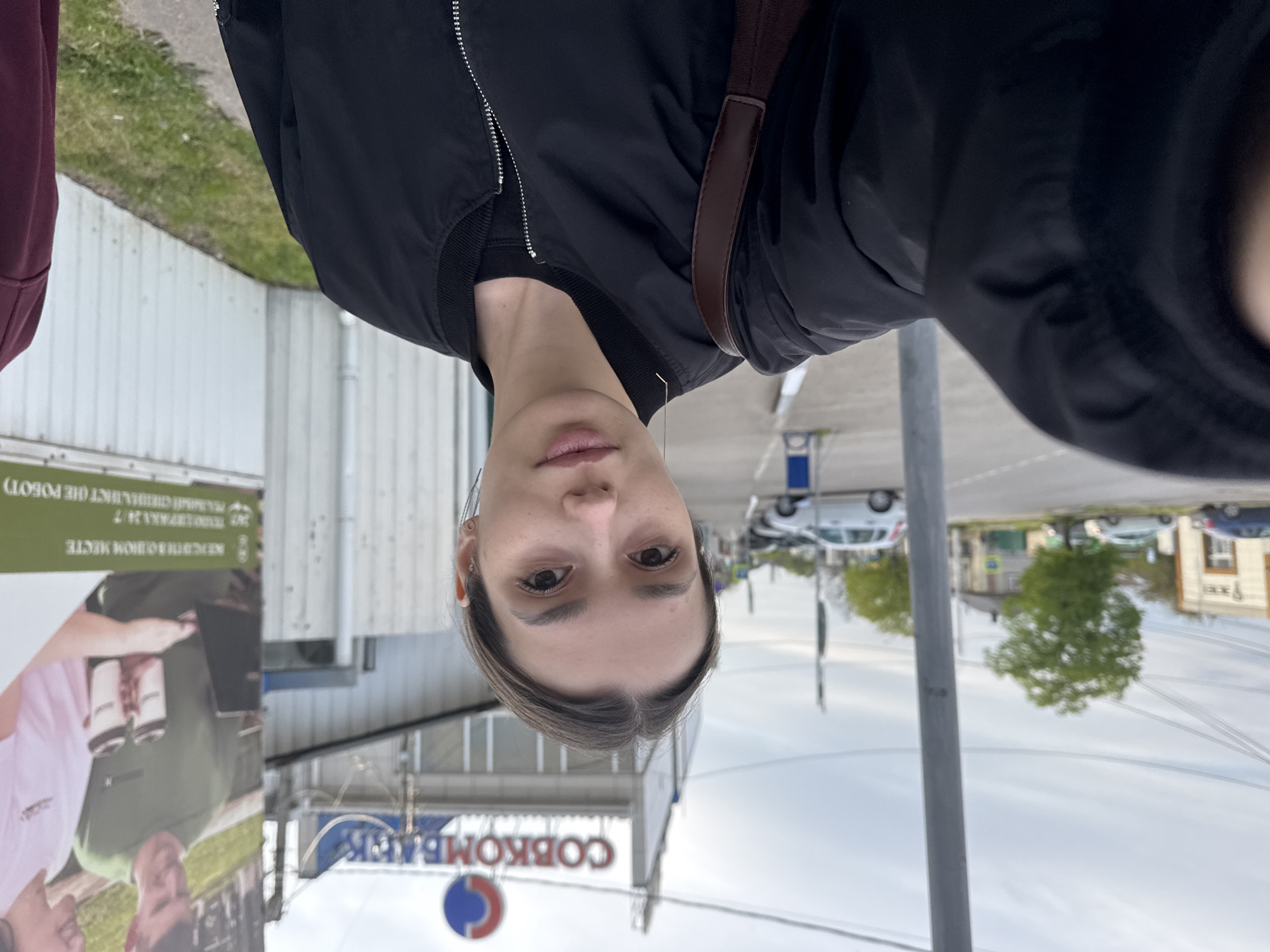 A selfie-style portrait captures a person with a neutral expression, looking directly at the camera. The subject has medium-length dark hair parted in the middle and is wearing a black bomber jacket over a dark top, with a brown bag strap visible over their shoulder.
The background reveals an urban street scene under an overcast sky, suggesting daytime. To the left, a building with a white corrugated metal facade is partially visible. In front of this building stands a large advertising billboard featuring a person holding canned beverages. Text on the billboard is in Cyrillic script and includes phrases like "ПИВНАЯ КАРТА 24/7" (Beer Map 24/7) and "КРУГЛОСУТОЧНОЕ МЕСТО" (Round-the-clock place).
In the mid-ground, a prominent sign with a blue and red circular logo is visible, bearing the Cyrillic text "СОВКОМБАНК" (Sovcombank), which is a Russian bank. To the right of the subject, a street with parked cars and distant trees can be seen. Overhead wires are also visible in the distance. The presence of Cyrillic text on both the billboard and the bank sign strongly indicates the location is a Slavic-speaking country, most likely Russia.