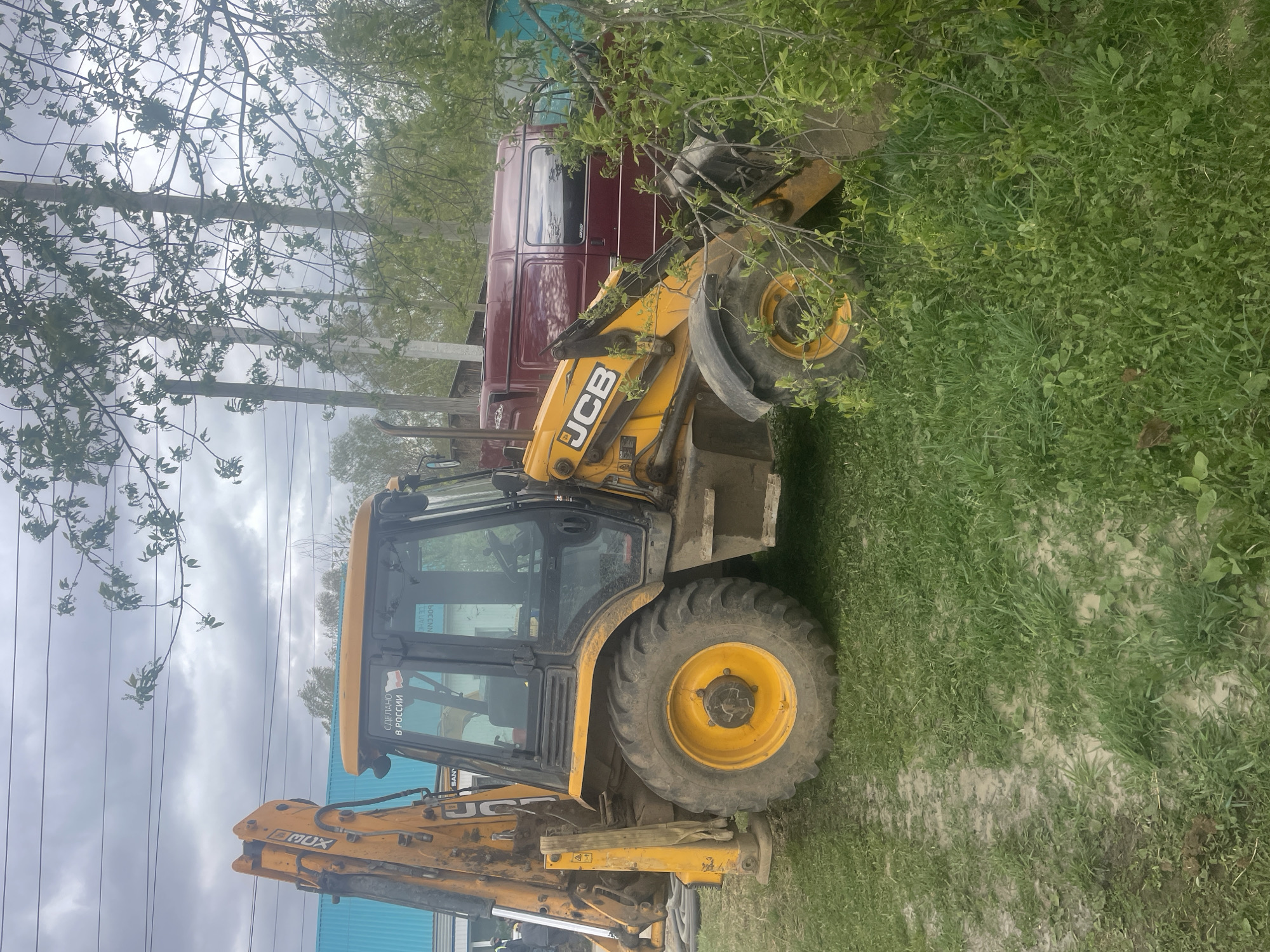 A yellow JCB backhoe loader is parked on a grassy area near a red truck in Поедуги, Russia. Overhead power lines and a cloudy sky are visible above the scene. The area has trees and vegetation.