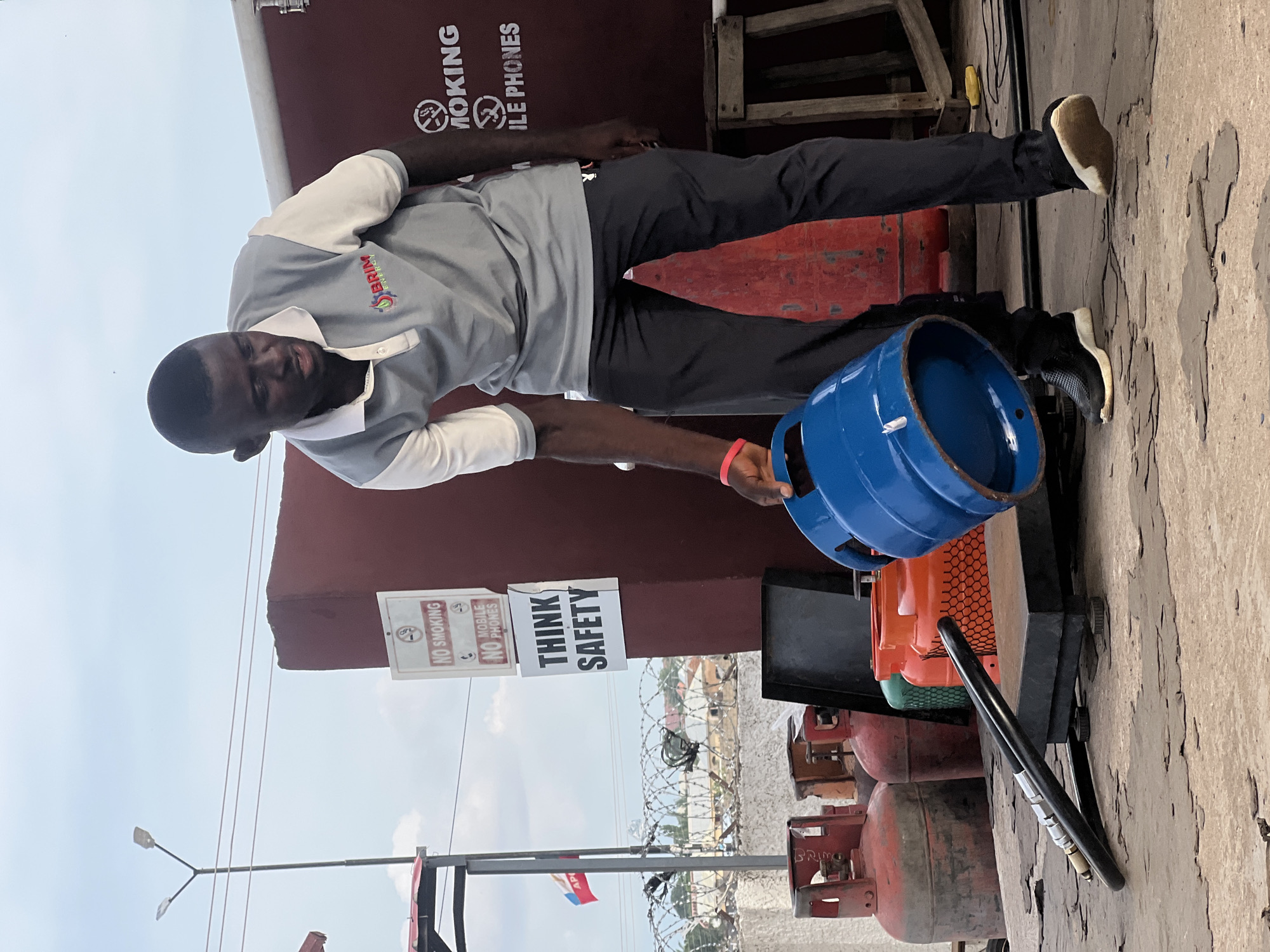 In Jalingo, Nigeria, a Black man wearing a grey and white polo shirt with a "BRIM ENERGY" logo and dark pants is holding a blue gas cylinder. He is standing on a rough, textured surface with several red and orange gas cylinders behind him. A black hose lies on the ground in front of him. A sign on the dark red wall behind the man reads "NO SMOKING" and "NO MOBILE PHONES", with another sign below it stating "THINK SAFETY". A street light and barbed wire fencing are visible in the background, suggesting an outdoor commercial or industrial area. The sky is overcast.