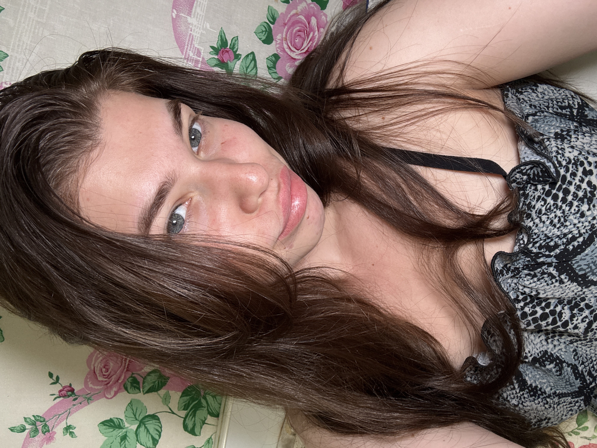 A young woman with fair skin, dark brown hair, and light blue eyes is lying on a floral patterned surface. She is wearing a grey and black animal print top with a black strap visible near her neckline. The background is a floral pattern of roses and green leaves. The location is not specified, nor can be derived, from the image.