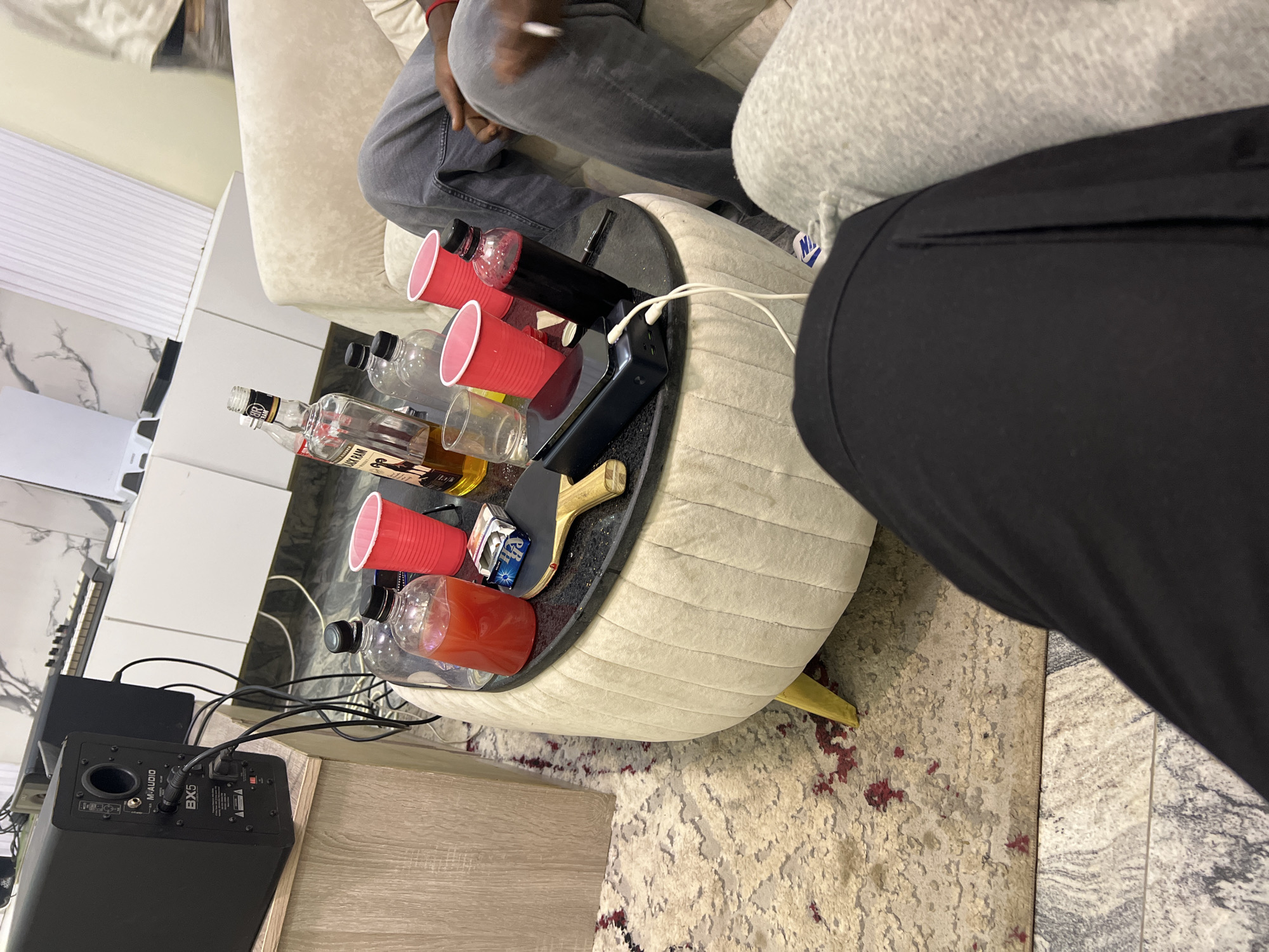 This media file captures an indoor scene, likely a living room or lounge area in Gwarinpa, Nigeria, characterized by a casual gathering. Two individuals are partially visible, seated on a light-colored, tufted sofa. One person wears grey jeans and a red wristband, holding a white object in their hand. The other person is seen wearing grey sweatpants with a visible Nike logo, under black pants.
At the center is a round, upholstered ottoman-style coffee table with a dark, reflective top surface and golden legs. The table is laden with various items: several red plastic cups (some empty, one containing a dark liquid, and another with a red/orange liquid), clear plastic bottles (one empty, one containing a dark liquid), and a glass liquor bottle labeled "OLD NICK RUM." Also on the table are a black power strip or charging station with multiple white cables plugged into it, a small wooden slingshot-like object, and a blue and white box.
To the left, a black M-AUDIO BX5 studio monitor speaker is visible on a light wood-grain surface, connected by several cables. Behind it, a musical keyboard or synthesizer can be partially seen. The flooring consists of white and grey patterned tiles, partially covered by a light-colored rug with maroon accents. White louvered blinds or shutters are visible in the background. The scene suggests a relaxed social setting, possibly an informal get-together.