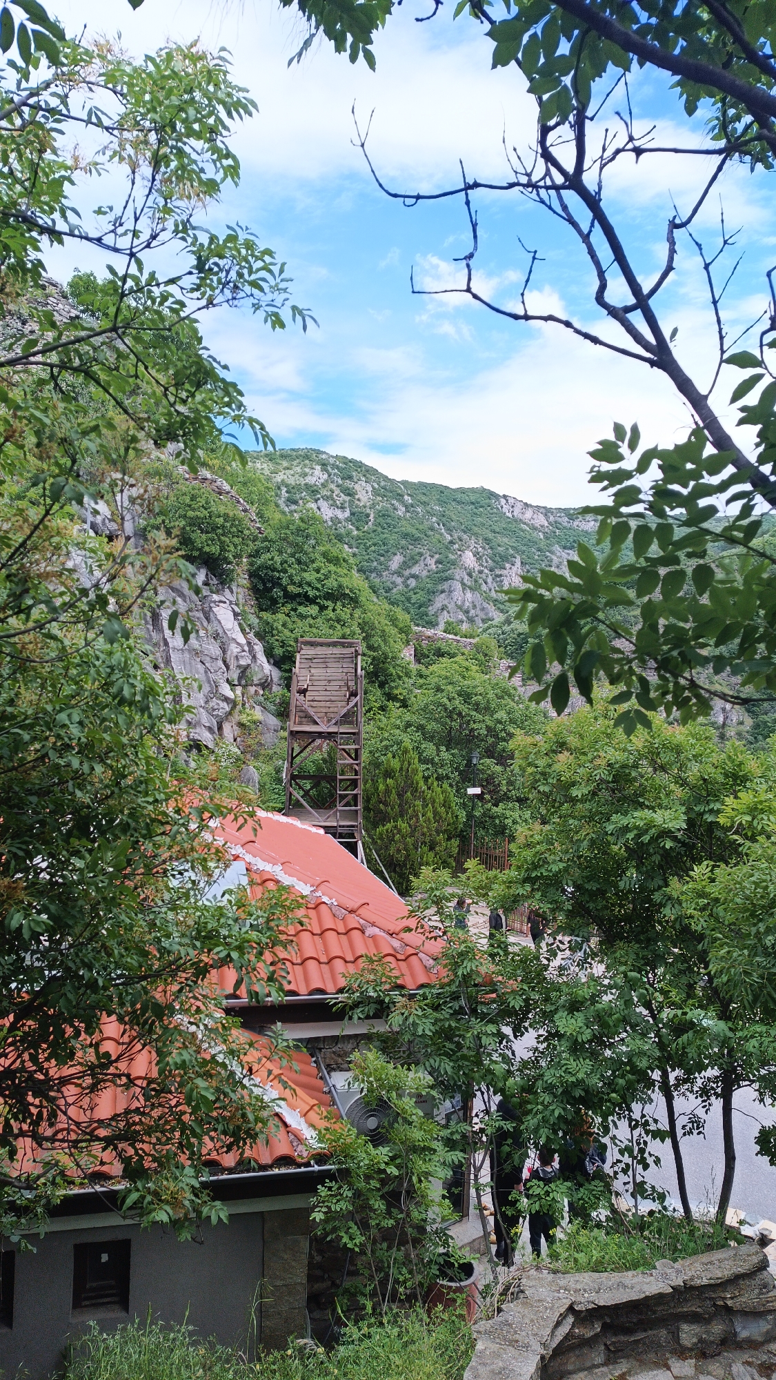 The image shows an exterior view in Lyaskovo, Bulgaria. A building with a red tile roof is partially visible. A wooden structure resembling a tower stands behind the roof. The background features a rocky, mountainous landscape with trees and vegetation. The sky is visible between trees with a blue sky and some white clouds. The foreground and middle ground are filled with trees, partially obscuring the view. Some people are standing on a roadside or sidewalk in the middle ground, behind the building with the red roof.