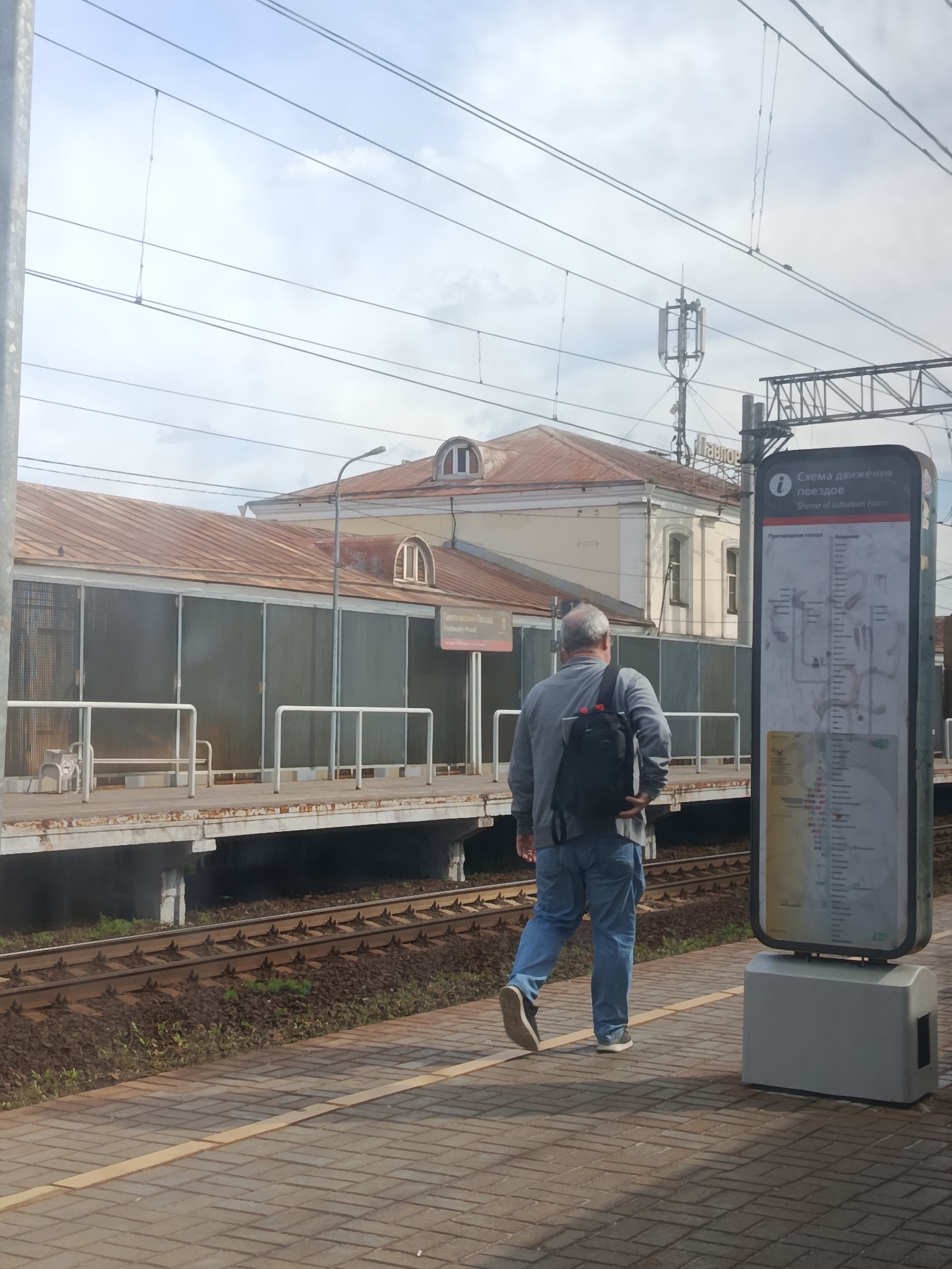 A man with gray hair, wearing a gray jacket and blue jeans, walks away from the camera on a train station platform. He carries a black backpack. To his right, a tall, slim, digital display board with a map of train routes stands on a gray base. The board has text in Russian and English, indicating it's a "Scheme of suburban trains." Behind the man and the display board is a train station building with a reddish-brown corrugated metal roof and beige walls. The building has arched windows and a covered platform area with metal railings and seating. Overhead, multiple power lines crisscross the sky, and a cellular tower is visible in the distance. The platform is paved with interlocking bricks, and a yellow line marks the edge. Train tracks are visible below the platform. A sign on the station building reads "Павловский Посад" (Pavlovsky Posad).