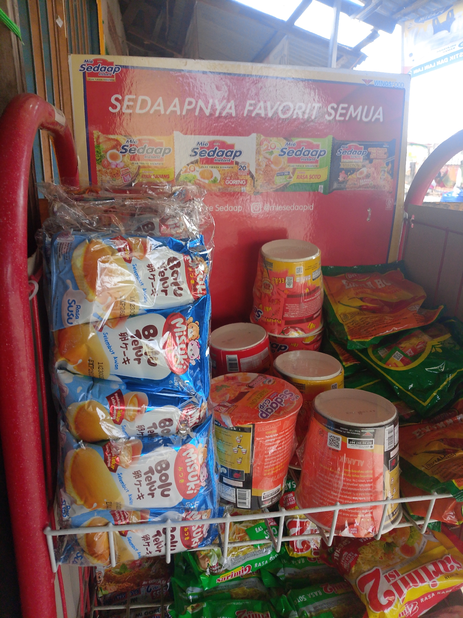 The image captures a close-up view of a street-side stall in Palembang, Indonesia, displaying an array of instant noodle products. The focal point is a prominent red and yellow display board featuring the "Mie Sedaap" brand, with the slogan "SEDAPNYA FAVORIT SEMUA" (Its Deliciousness is Everyone's Favorite). Below this board, several stacks of "Mie Sedaap" instant noodle cups and packets are arranged, showcasing various flavors such as Ayam Bawang (Garlic Chicken) and Rasa Soto (Soto Flavor).
To the left of the display board, a collection of individually wrapped "Bolu Telur Misuki" (Egg Cake) products are neatly stacked in a clear plastic wrapper. These cakes are depicted with an image of a sliced cake showing a yellow filling and a fluffy texture, and the packaging features both Indonesian and Japanese text.
The stall itself appears to be made of simple materials, with a white wire shelving unit holding some of the noodle products. In the background, the environment suggests an outdoor market setting, with hints of corrugated metal roofing and other goods visible. The lighting indicates it is daytime, with sunlight filtering in. No people are directly visible in the frame, but the presence of the merchandise implies a vendor's stall. The overall impression is that of a typical small business operation in a local Indonesian market.