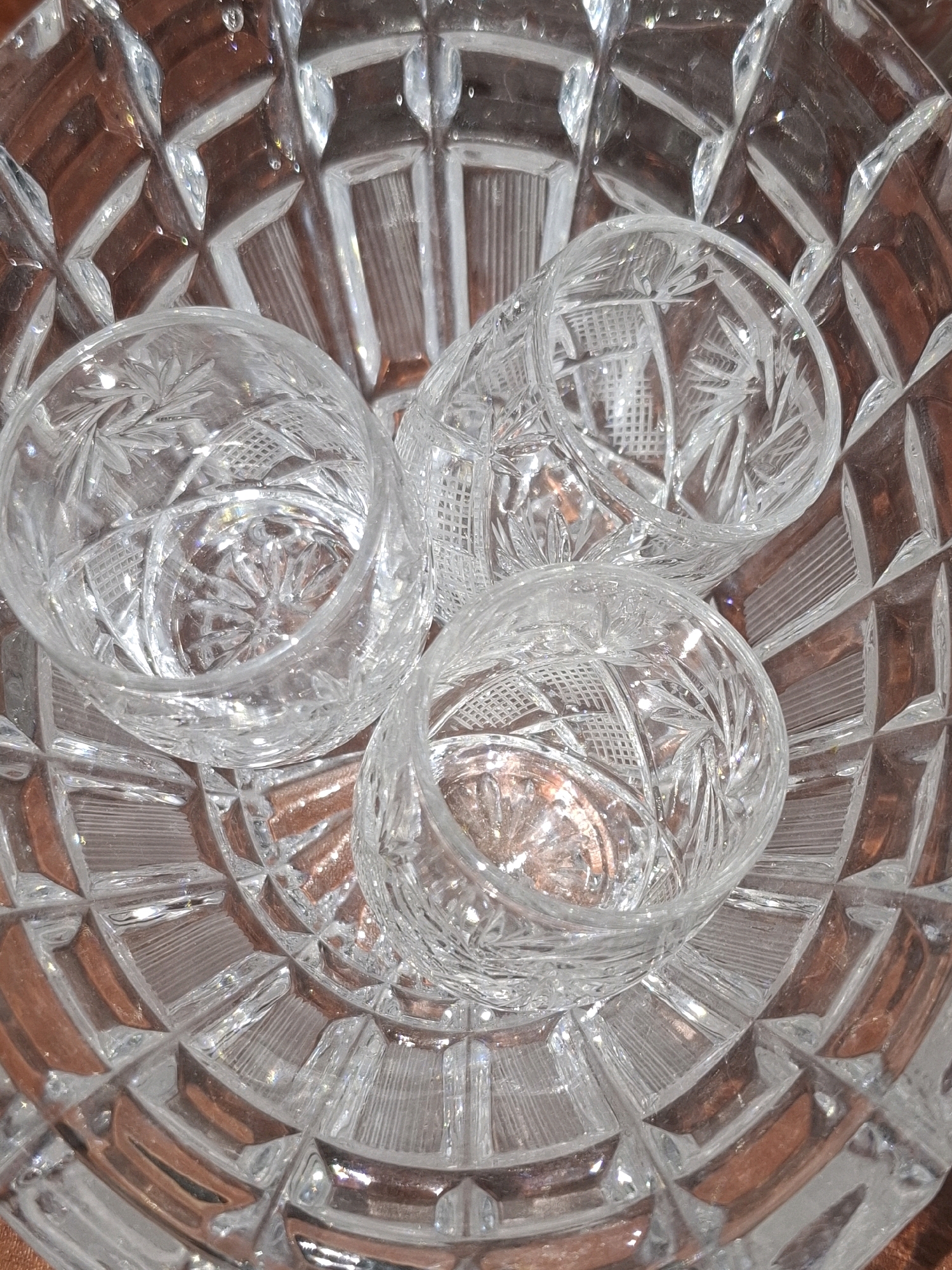 The image shows a close-up, top-down view of three small, clear, cut-glass shot glasses nestled within a larger, intricately patterned glass bowl. The shot glasses are arranged in a triangular formation, with one at the top and two below. Each glass has a cut-glass pattern, featuring starburst designs and diamond-shaped cuts. The large bowl also exhibits a detailed geometric pattern, with alternating facets and ribbed sections. The background appears to be a reddish-brown surface, possibly wood or fabric, visible through the transparent glass. The lighting creates numerous reflections and highlights on the glass surfaces, enhancing their sparkle and depth. The image captures the texture and clarity of the glass objects. There is no indication of any specific location in Almaty, Kazakhstan, or any environmental context beyond the objects themselves.
