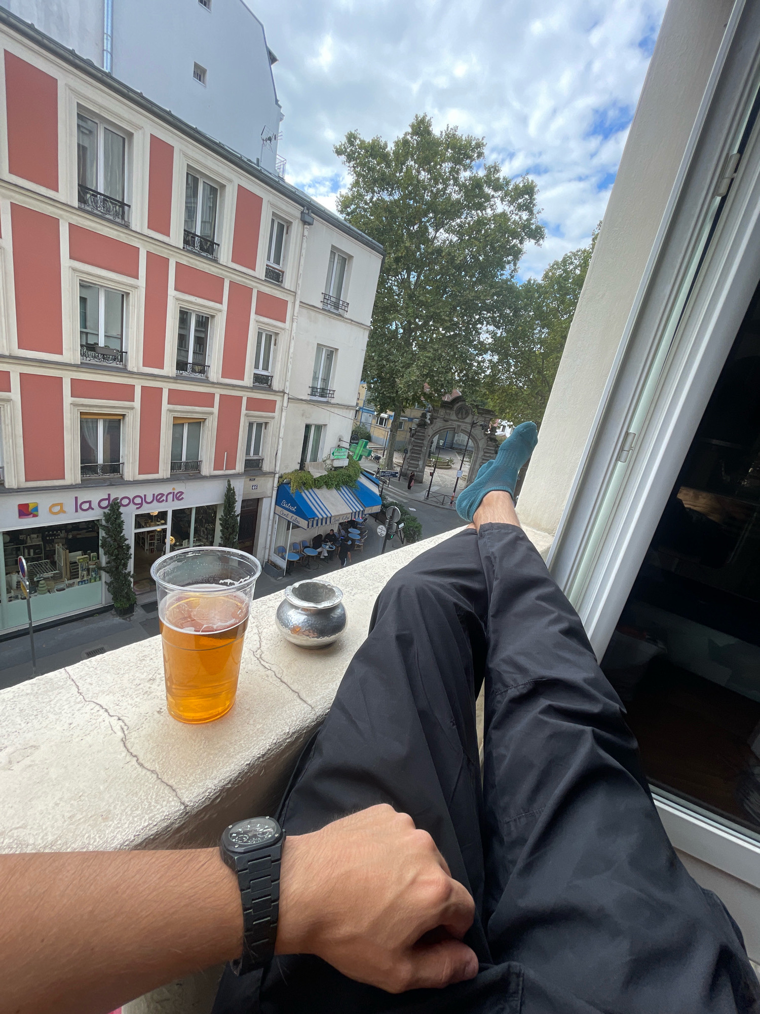 A person's legs, clad in black trousers and blue socks, are propped up on a balcony railing. A plastic cup of amber liquid, presumably beer, sits on the railing next to a small metal ashtray. In the foreground, a black digital watch is visible on the person's wrist. The background shows a European city street scene with buildings, trees, and awnings. The buildings have a distinctive architectural style with red and white facades, suggesting a location in Paris, France. The sky is partly cloudy.