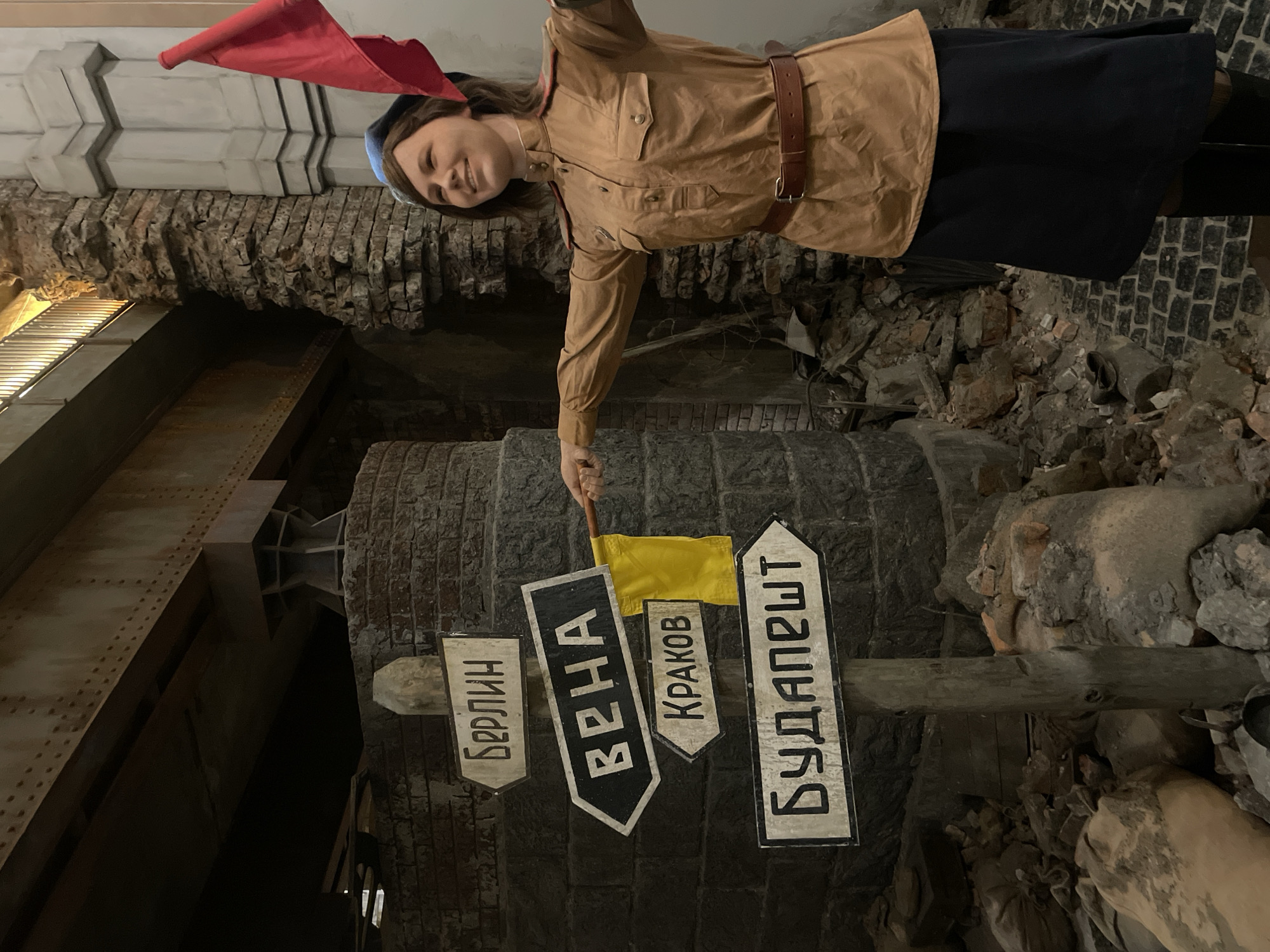 A woman dressed in a khaki Soviet-era military uniform with a red flag is standing in what appears to be a staged environment. The scene is located in the city of Moscow, Russia. Behind her are signs pointing towards cities such as Berlin, Vienna, Krakow, and Budapest. The environment is made to look like a war-torn area, with rubble and faux brickwork.