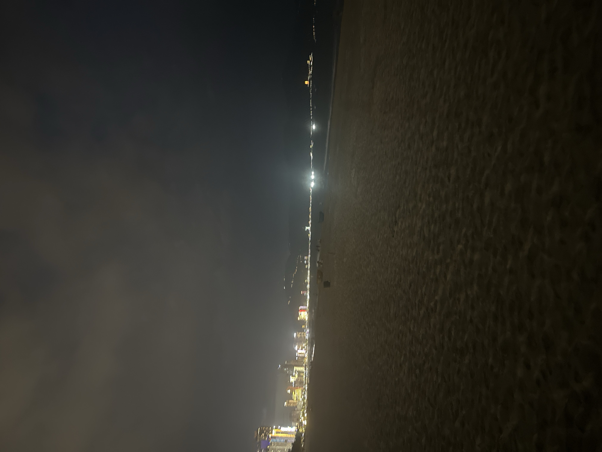 The image shows a nighttime view of a beach in Da Nang, Vietnam, with the city skyline illuminated in the background. The foreground is dominated by the dark sand of the beach, with subtle ripples indicating texture. The middle ground features the dark, calm water of the sea meeting the shore. Scattered on the beach are a few small, dark objects, possibly trash cans or beach chairs, and a couple of indistinct figures are visible walking on the sand near the water's edge.
The background is a vibrant display of the Da Nang cityscape. Tall buildings, lit with a variety of colorful lights, stretch across the horizon. Yellow, white, and red lights are particularly noticeable, suggesting hotels, apartments, and commercial buildings. Further in the distance, lights fade into the darkness, hinting at the extent of the city. The sky above is dark and cloudy, with some of the city lights reflecting faintly on the cloud cover. The overall scene conveys a sense of a lively coastal city at night.