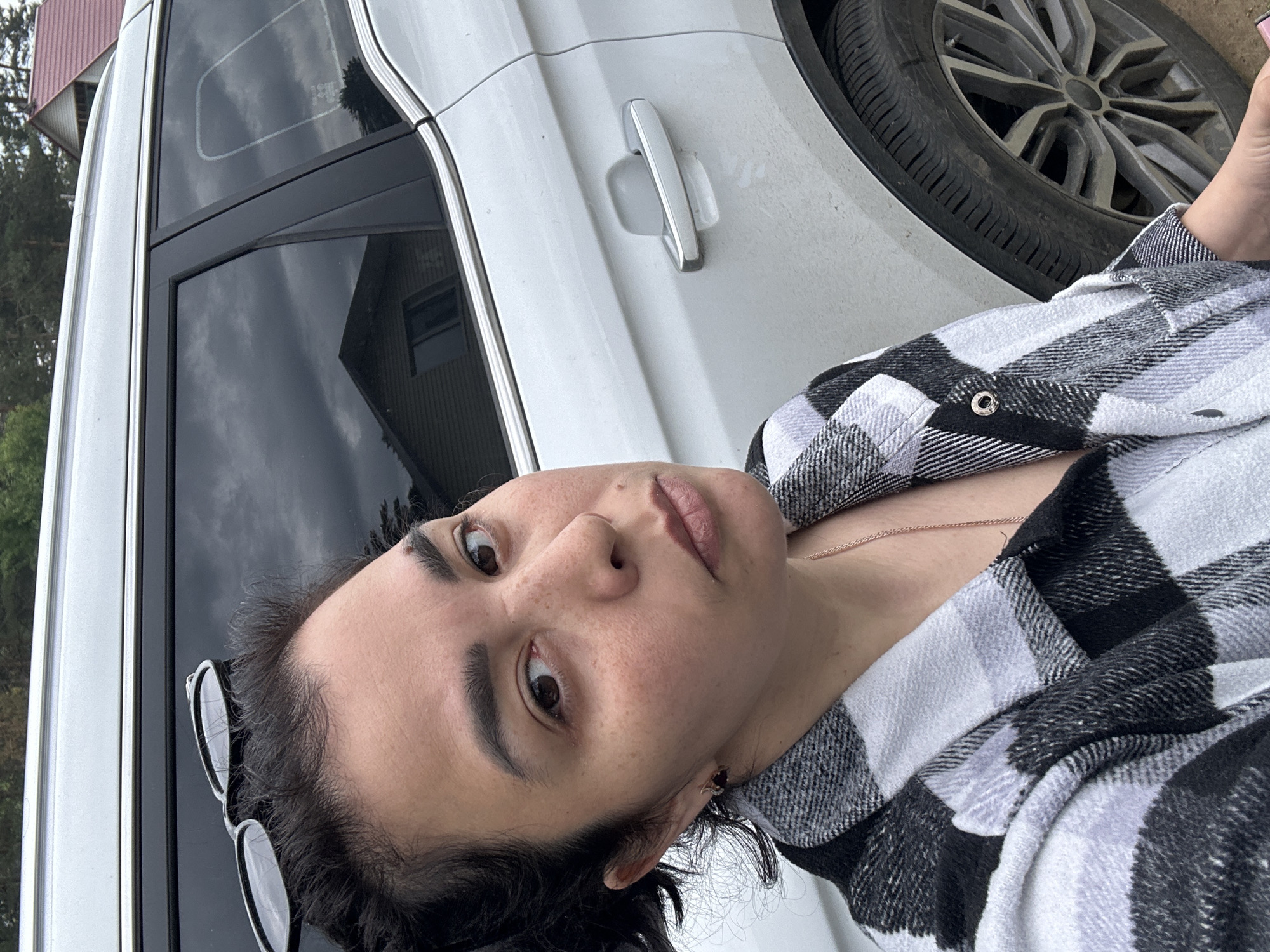 A close-up, low-angle shot, rotated 90 degrees clockwise, features a woman taking a selfie or being photographed outdoors next to a white vehicle. The woman, with dark hair and dark eyes, has a neutral to slightly serious expression and is looking directly at the camera. She wears a black and white plaid flannel shirt, and a thin, gold-colored necklace is visible around her neck. Black sunglasses are perched on her head.
To her right (top of the rotated image) is a white car, likely an SUV, with a visible door handle and tinted windows. The car's front or rear wheel and tire are also partially visible. Reflections in the car windows show a cloudy sky, trees, and parts of what appears to be a house or building with a reddish roof, suggesting a suburban or rural outdoor setting. The lighting appears soft and diffused, consistent with an overcast day. No discernible text is visible.
