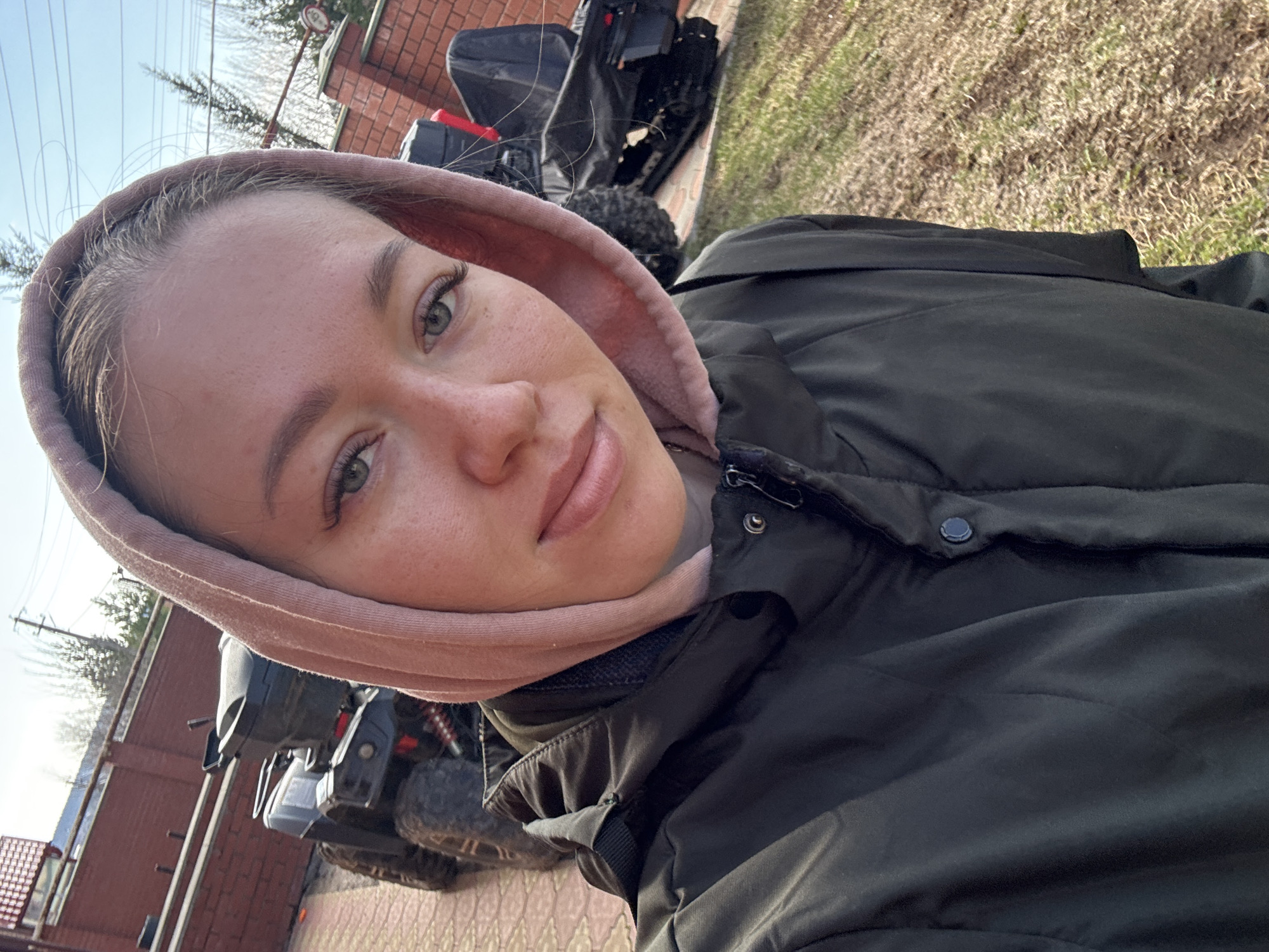 The image is a vertical selfie capturing a young woman outdoors on a clear day. She is seen in a close-up shot, with her head slightly tilted and her gaze directed towards the camera. She has light-colored hair, green or hazel eyes with visible eyelashes, and light freckles across her nose and upper cheeks. Her expression is calm, with a subtle hint of a smile.
She is wearing a light pink hooded sweatshirt, which frames her face, layered under a dark, possibly black or dark green, padded jacket. The jacket has visible seams and a button on the front.
The background suggests an outdoor, possibly rural or semi-rural setting. Partially visible behind her, to the left and upper right, is a dark-colored All-Terrain Vehicle (ATV) or quad bike, featuring robust tires and red suspension parts. A reddish-brown brick or wooden fence or wall is present in the mid-ground. The ground is covered with sparse vegetation, primarily dried, light brown grass mixed with some greener patches. The sky above is bright and clear, with several power lines or wires visible against it. The lighting indicates it is daytime, likely sunny. The woman appears relaxed. There is no visible text in the image. The scene aligns with the provided location context of a farm ("Ферма") in Russia.