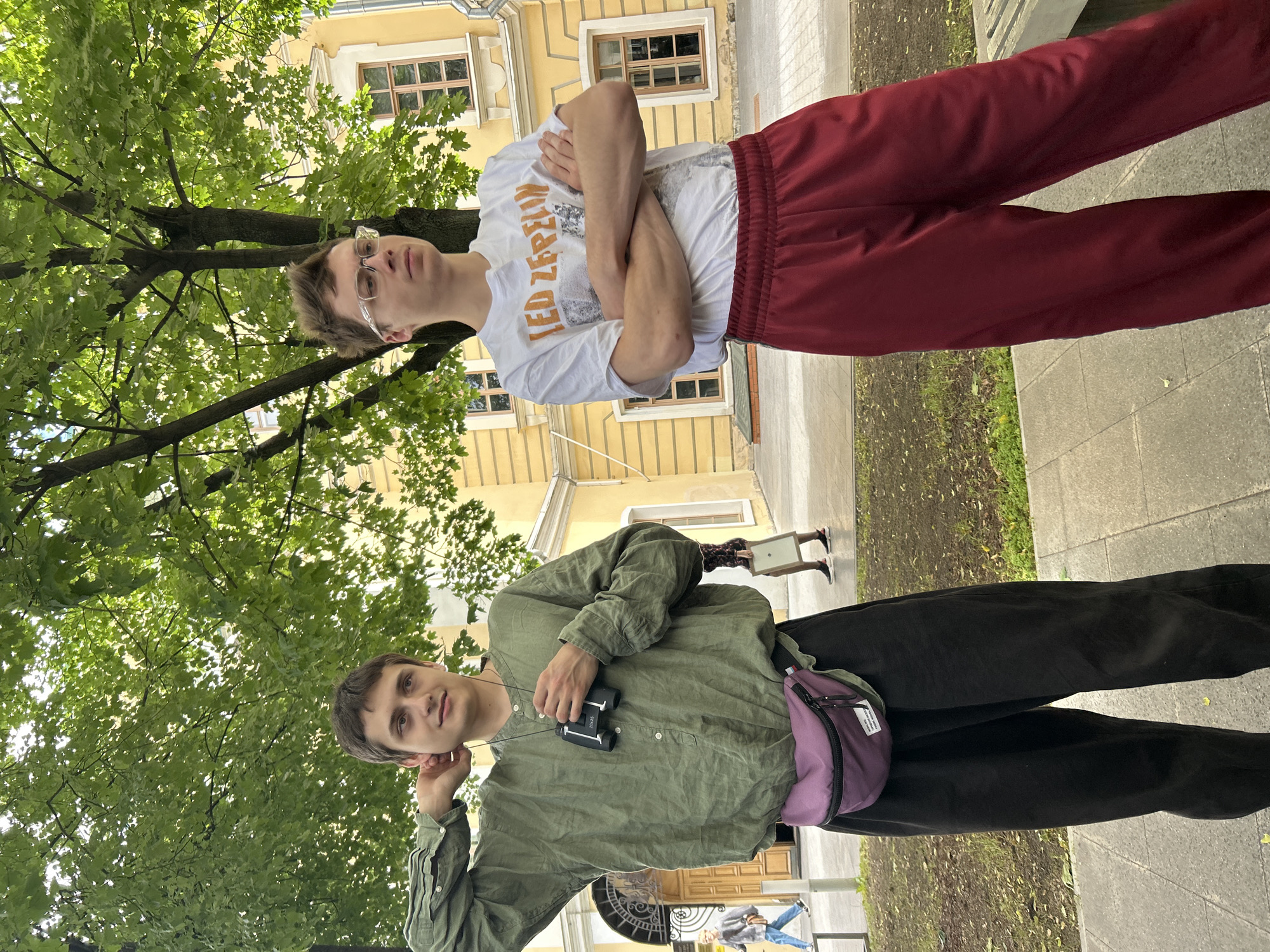 Two young men are standing outdoors in front of a yellow building with a tree overhead. The man on the left is wearing a loose-fitting olive green shirt with a purple fanny pack around his waist. He is holding a pair of binoculars. The man on the right is wearing a white t-shirt with a "Led Zeppelin" graphic and dark red sweatpants. He has his arms crossed. There are paved walkways and some greenery visible. The lighting suggests it is daytime.