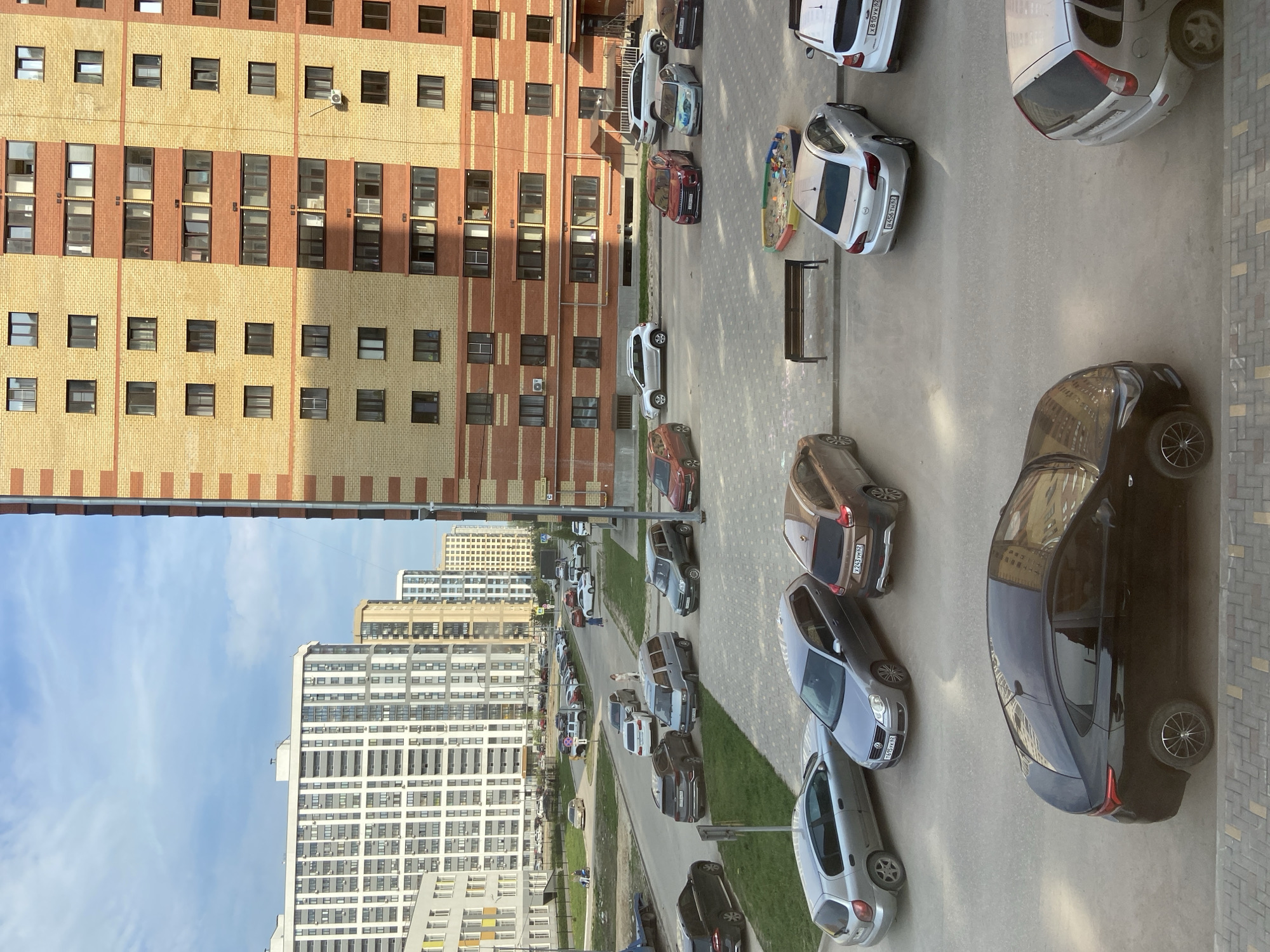 Here's a detailed description of the image:
**Setting:** The image appears to be taken from a high vantage point, likely from an upper floor of a building in Дядьково, Russia. The buildings in the foreground and background suggest a dense urban residential area. The sky is partially visible with a blue hue indicating a clear day.
**Buildings:** There's a large, multi-story apartment building with a brick facade consisting of alternating bands of yellow and red brick. Other residential high-rise buildings are visible in the distance, colored primarily in white.
**Transportation:** A street runs between the buildings, packed with parked cars. The street is paved in grey tarmac.
**Vehicles:** Numerous cars are parked along the sides of the road, facing different directions. A silver-gray sedan stands out in the foreground, partially off-frame. Car colors include silver, red, brown, gray, and white.
**Other Objects:** A small children's playground with a sandpit is visible next to one of the buildings. A metal bench is seen next to the playground. Patches of grass are noticeable between parked cars along the side of the road.
**People:** No people are clearly visible.