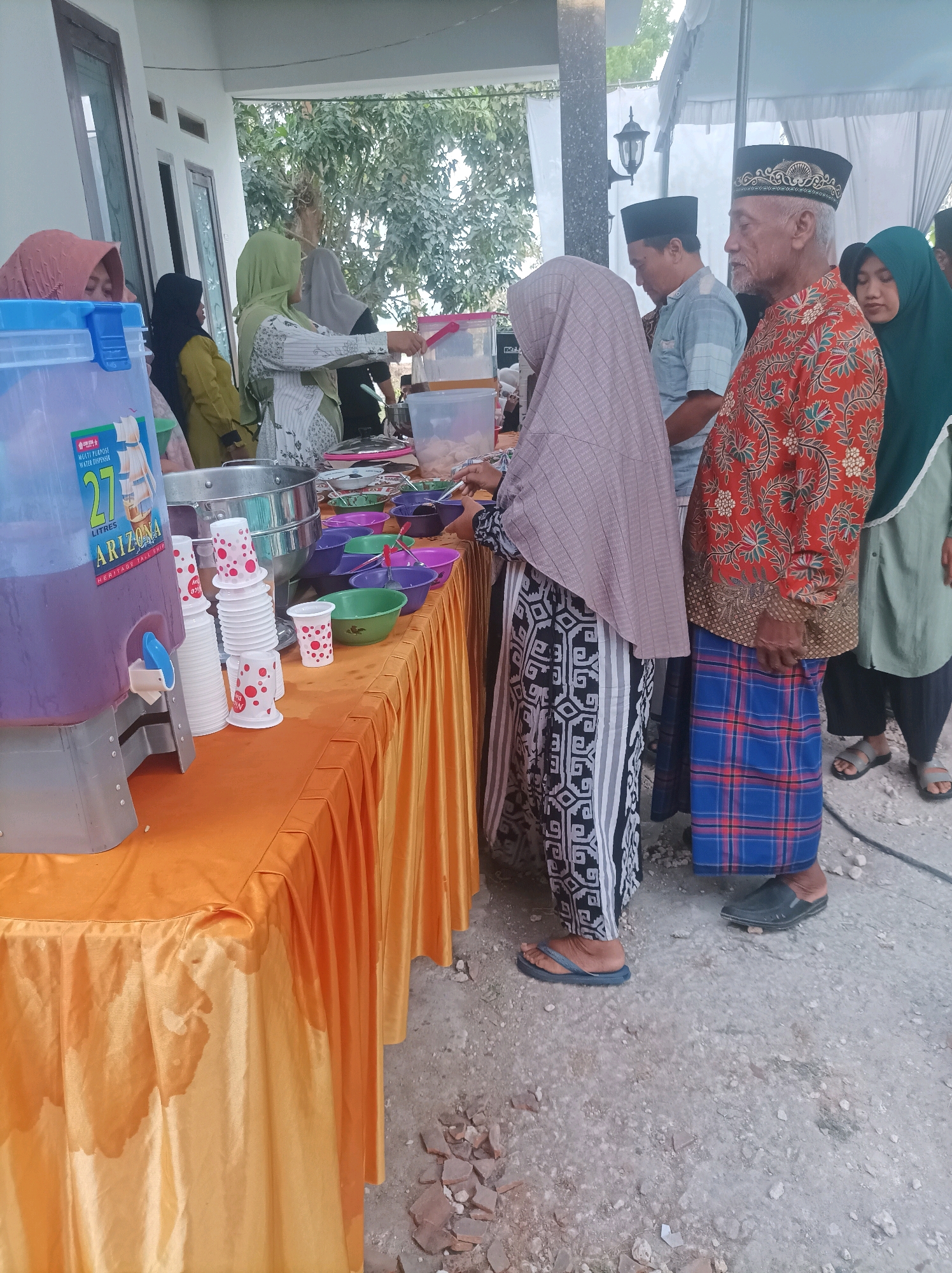 In Canga'an, Indonesia, an outdoor event is taking place, possibly a gathering or celebration, with food and drink being served buffet-style. A prominent large dispenser filled with purple liquid, labeled "ARIZONA 27 LITRES," stands on an orange-covered table. Next to it, stacks of white paper cups with red polka dots are arranged. Various serving bowls and utensils are spread across the table, suggesting a meal is being offered.
Several individuals are present. On the left, a woman wearing a light pink hijab is partially visible. To her right, another woman in a yellow hijab stands near the dispenser. Further down the table, a person in a green hijab is engaged with the serving area. Towards the right side of the image, an elderly man with a white beard, wearing a traditional patterned red shirt and a peci (a cap), is talking with another man in a light blue shirt. Behind them, a woman in a dark green hijab is also present.
The setting appears to be a somewhat informal outdoor space, with a covered area in the background and uneven ground with some debris. The overall scene depicts a moment of communal dining or refreshment.