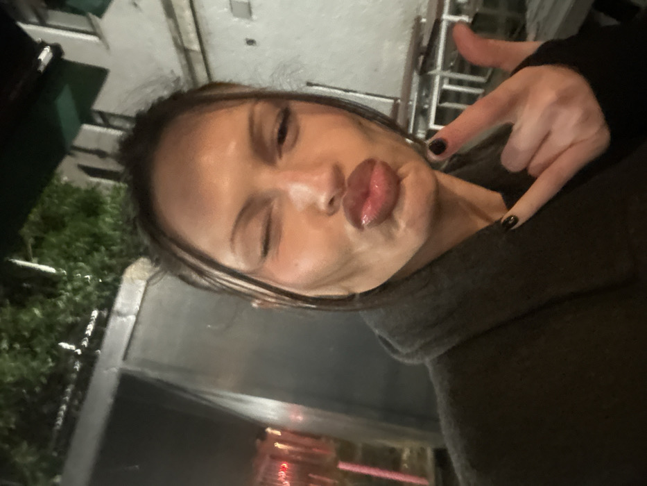 A woman, with dark hair pulled back, puckers her lips for a selfie. She winks one eye. Her dark nails are visible as she makes a rock-and-roll hand gesture. Part of a building or structure is visible to the left of her face, partially obscured by foliage. There are reflections in the lower section of the frame. The overall lighting is low. She wears a dark, heavy sweater.