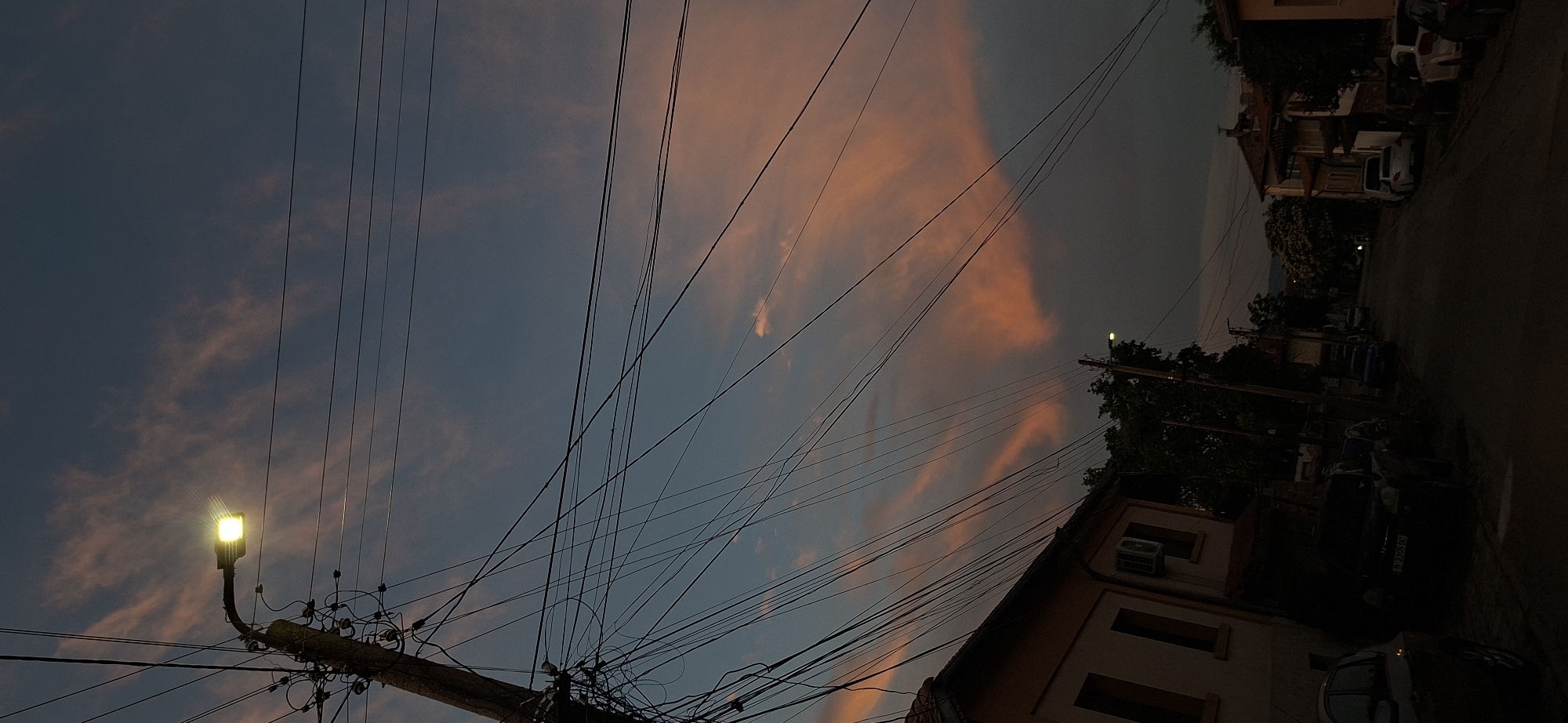 The image depicts a street scene at dusk in Lyaskovets, Bulgaria. The sky is a dusky blue with streaks of pink and orange clouds, illuminated by a bright street lamp. A wooden utility pole, heavily laden with tangled wires, stands on the left side of the frame, casting shadows. Adjacent to the pole is the side of a building with a light-colored facade, featuring windows and an air conditioning unit. The street is lined with parked cars, some partially visible in the dim light. Further down the street, more buildings and trees are visible, with a hint of distant hills. The overall atmosphere is somber and quiet, characteristic of the end of the day in a residential area.