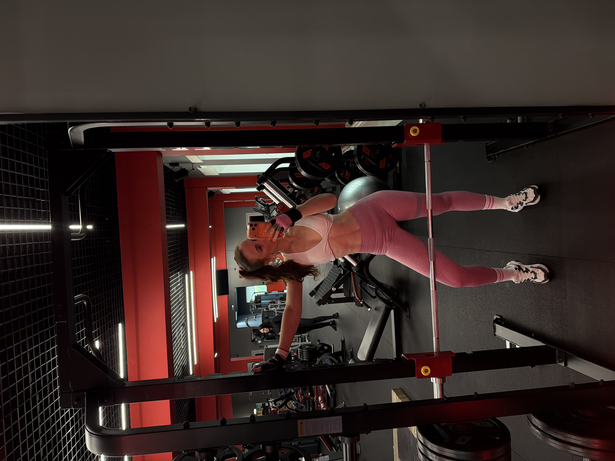 The image depicts a young woman taking a mirror selfie inside a modern gym in Birobidzhan, Russia. The woman, with long brown hair, is wearing a light pink sports bra, matching pink textured leggings, white athletic socks with pink stripes, and white and black athletic shoes. She holds an orange-colored smartphone with a pink case in her left hand, angled towards the mirror to capture her reflection. Her right hand, adorned with a black and pink fingerless glove, rests on a piece of black gym equipment, likely part of a smith machine or squat rack. She has a pleasant expression with a slight smile.
The gym features a striking interior design with red walls and black grid-patterned panels. Bright, elongated white strip lights illuminate the space. In the background, various pieces of gym equipment are visible, including weight plates marked "25" and "32," and a grey exercise ball. Another person, dressed in dark clothing, is faintly reflected in the mirror deeper within the gym, suggesting an active environment. A small warning or instructional label is visible on the black gym equipment in the mid-ground. The floor is dark, possibly rubberized gym flooring. The overall setting is well-lit, suggesting either daytime or evening indoor activity.