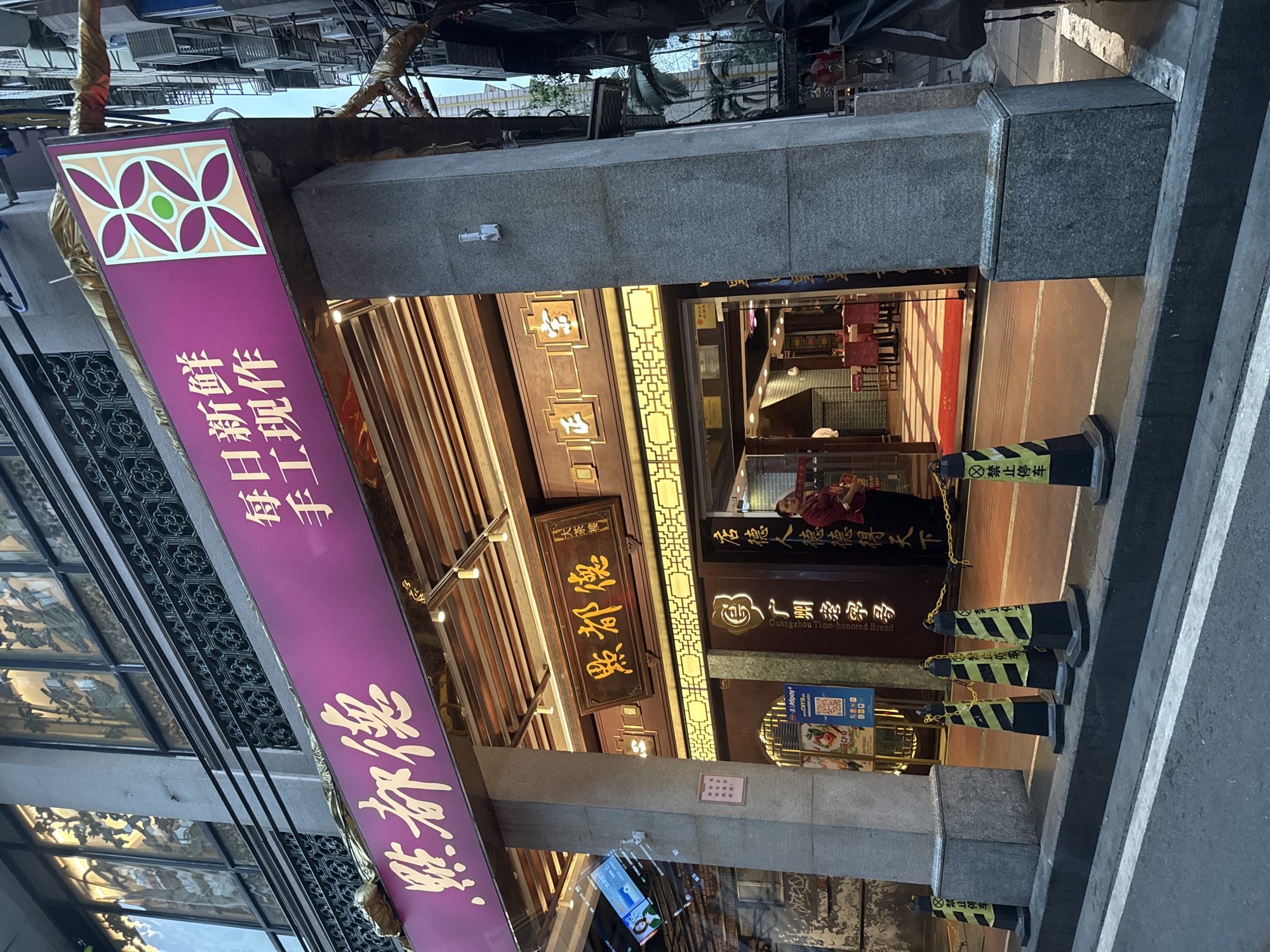 The image captures the facade of a restaurant named "Dian Du De" (點都德), with text in both English and Chinese indicating that it's a "Guangzhou Time-honored Brand". A purple banner to the left features the restaurant's name in large Chinese characters along with smaller text that translates to "Fresh daily, handmade". Inside, there is evidence of a traditional Chinese interior design with wooden elements. A woman in a red shirt can be seen inside the restaurant. Safety cones with yellow and black stripes line the edge of the sidewalk in front of the restaurant. The image appears to be taken in the Liwan District of China.
