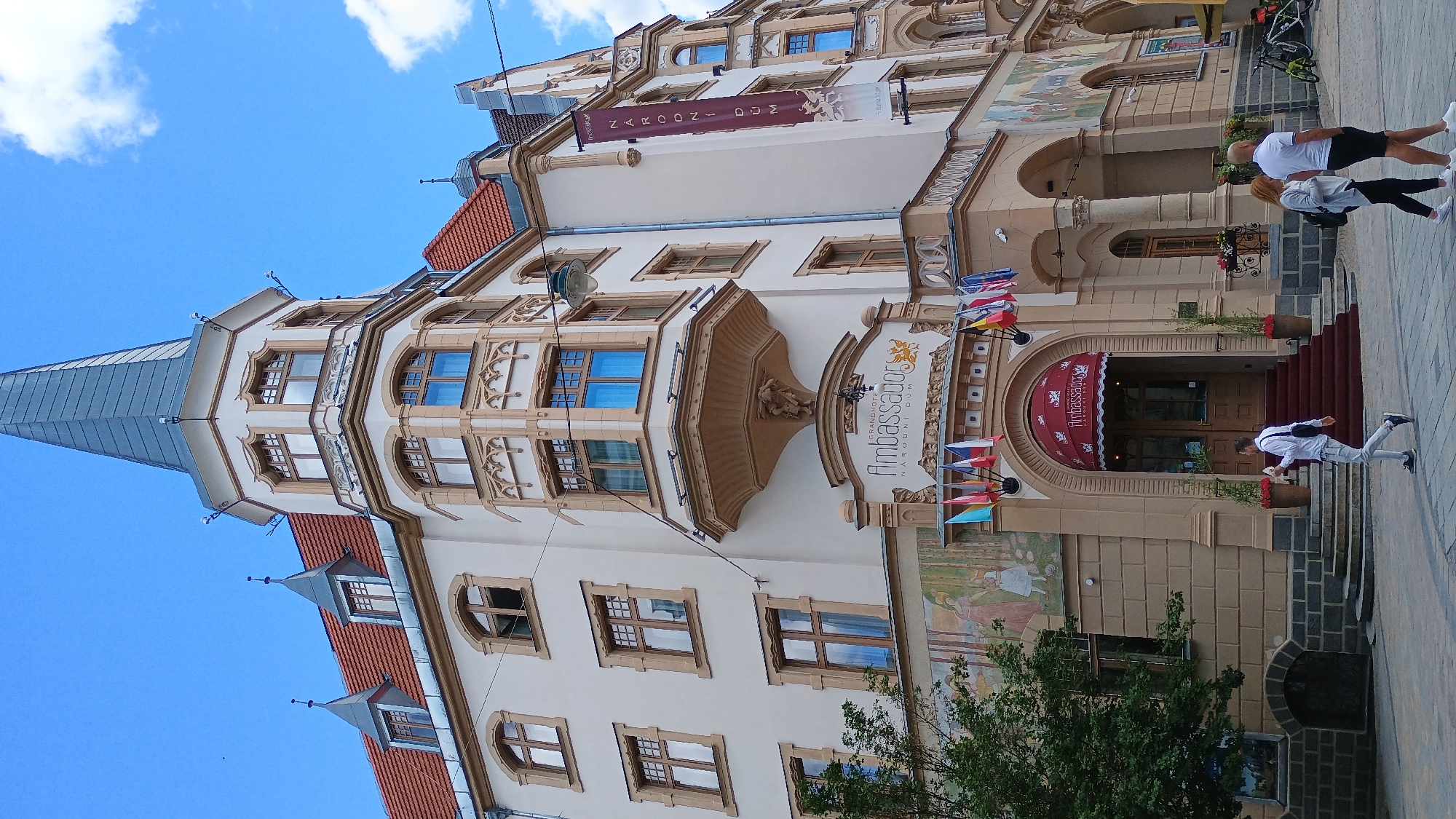 The image shows the Grandhotel Ambassador, Narodni Dum, located in Karlovy Vary, Czechia. The hotel is a multi-story building with an ornate facade, featuring bay windows, decorative elements, and a prominent spire. A red awning with the hotel's name is visible above the entrance, with a set of red stairs leading up to double doors. Several small flags are displayed above the entrance. In front of the hotel, a man in a white shirt and light-colored pants is walking, holding a white object. To the right of the entrance, a couple is standing and looking in the same direction. The background shows a clear blue sky with some clouds. The scene appears to be a street with some greenery and bicycles visible in the distance.