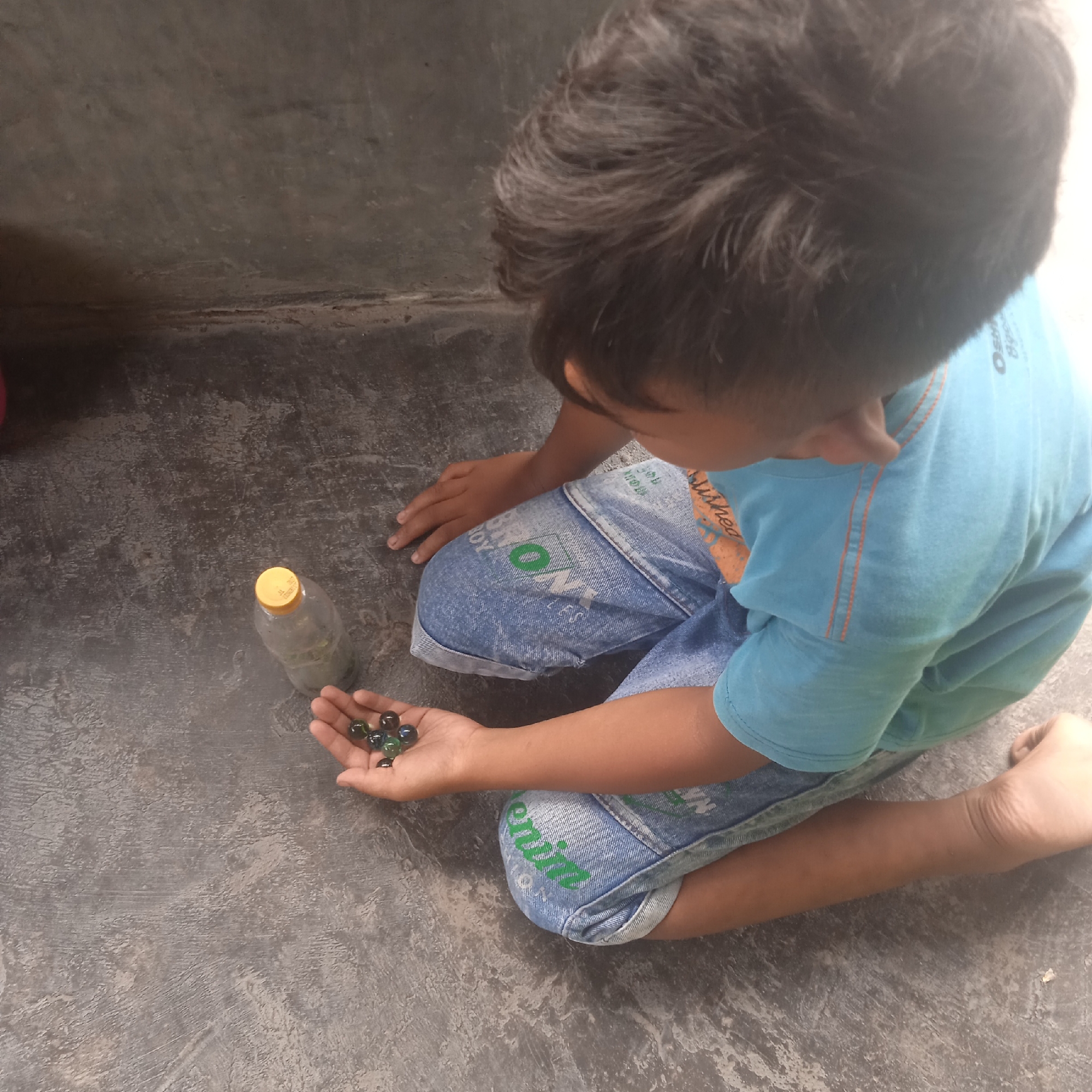 In Situterate, Indonesia, a young boy kneels on a grey, textured surface, likely a concrete ground. He is wearing a blue t-shirt with orange trim and denim shorts. In his right hand, he holds several marbles, including some that appear to be green and dark-colored, with a few reflecting light. Beside him is a clear plastic bottle with a yellow cap, containing a murky liquid. The boy is looking down at the marbles in his hand, engaged in play. The background consists of a plain, grey wall.