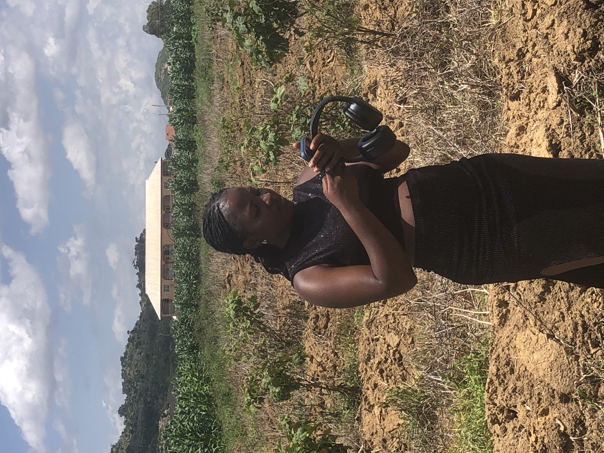A Black female stands in a field of dry earth and sparse vegetation, holding a mobile phone and a pair of black over-ear headphones. She wears a black sleeveless, sequined crop top and a black skirt with a thigh-high slit. The background features a distant building with a light-colored roof, behind a field of green crops, possibly corn. Hills covered in greenery are visible in the far distance under a partly cloudy sky. The scene appears to be in a rural or semi-rural area, potentially on the outskirts of Gwafan, Nigeria.
