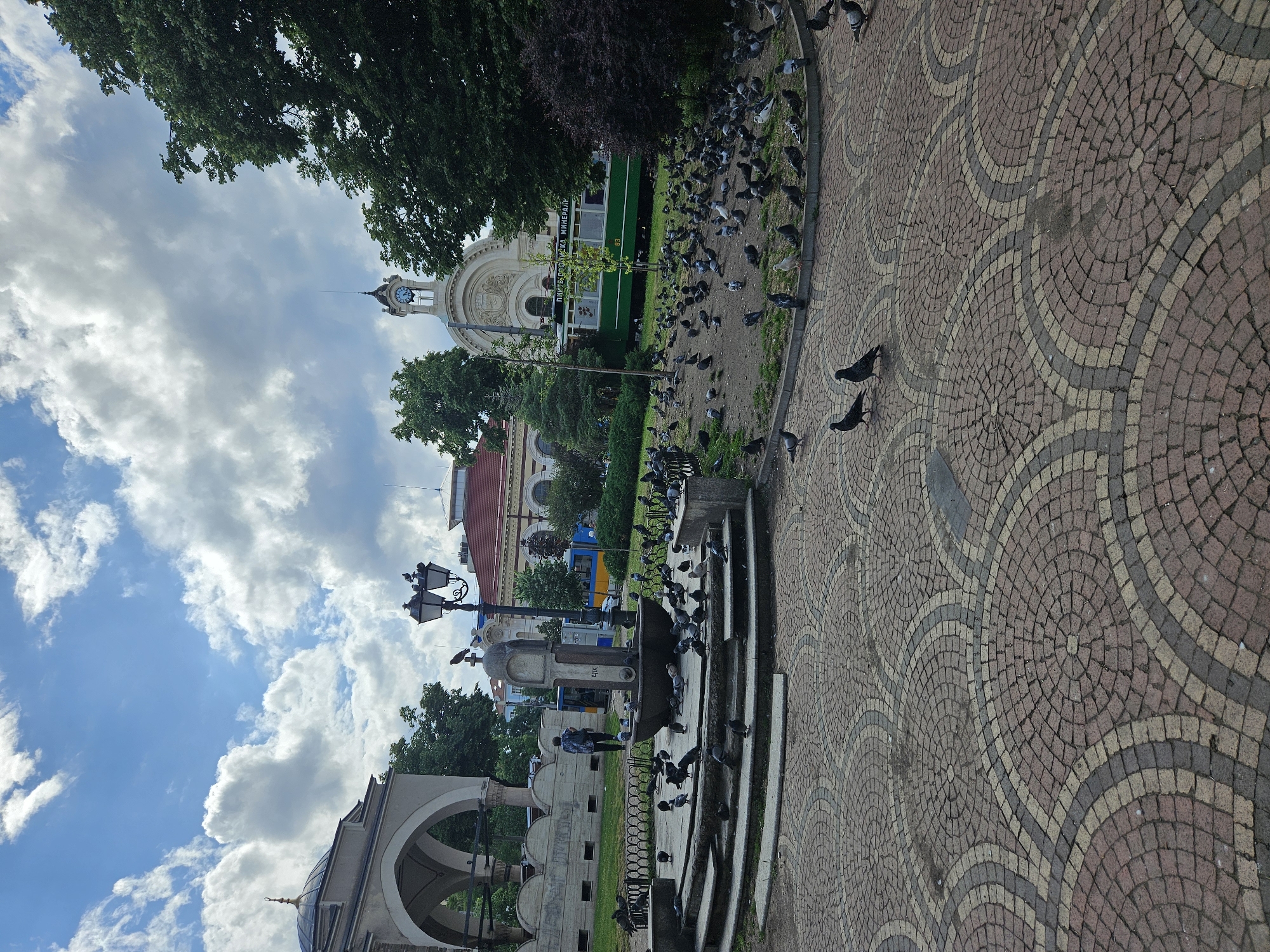 This image depicts a lively urban square in Sofia, Bulgaria, characterized by prominent historical architecture and a bustling flock of pigeons.
In the foreground, a large expanse of patterned cobblestone pavement covers the right side of the frame, featuring intricate wave-like designs in shades of red, beige, and grey. Numerous pigeons are scattered across this pavement and the adjacent grassy areas, creating a dynamic scene. Many more pigeons congregate around a multi-tiered stone fountain or monument in the mid-ground, with a person walking nearby.
To the left, the distinctive dome and arched entrance of the Banya Bashi Mosque are partially visible. Dominating the background is the grand, ornate facade of the former Sofia Central Mineral Bath, now housing the Regional History Museum, complete with a prominent clock tower. A green tram or bus is parked in front of this building; visible text on its side reads "ПИТЕЙНА РАЗЛИВНА МИНЕРАЛНА ВОДА" (Drinking Bottling Mineral Water) and the number "03". This indicates a mineral water vending point, a common feature in the area.
Lush green trees frame the top and middle sections of the image. The scene is illuminated by bright daylight under a partly cloudy sky, with patches of blue visible between fluffy white clouds. Traditional black street lamps are also part of the urban landscape. The overall atmosphere suggests a vibrant public space where historical architecture meets everyday city life and nature.