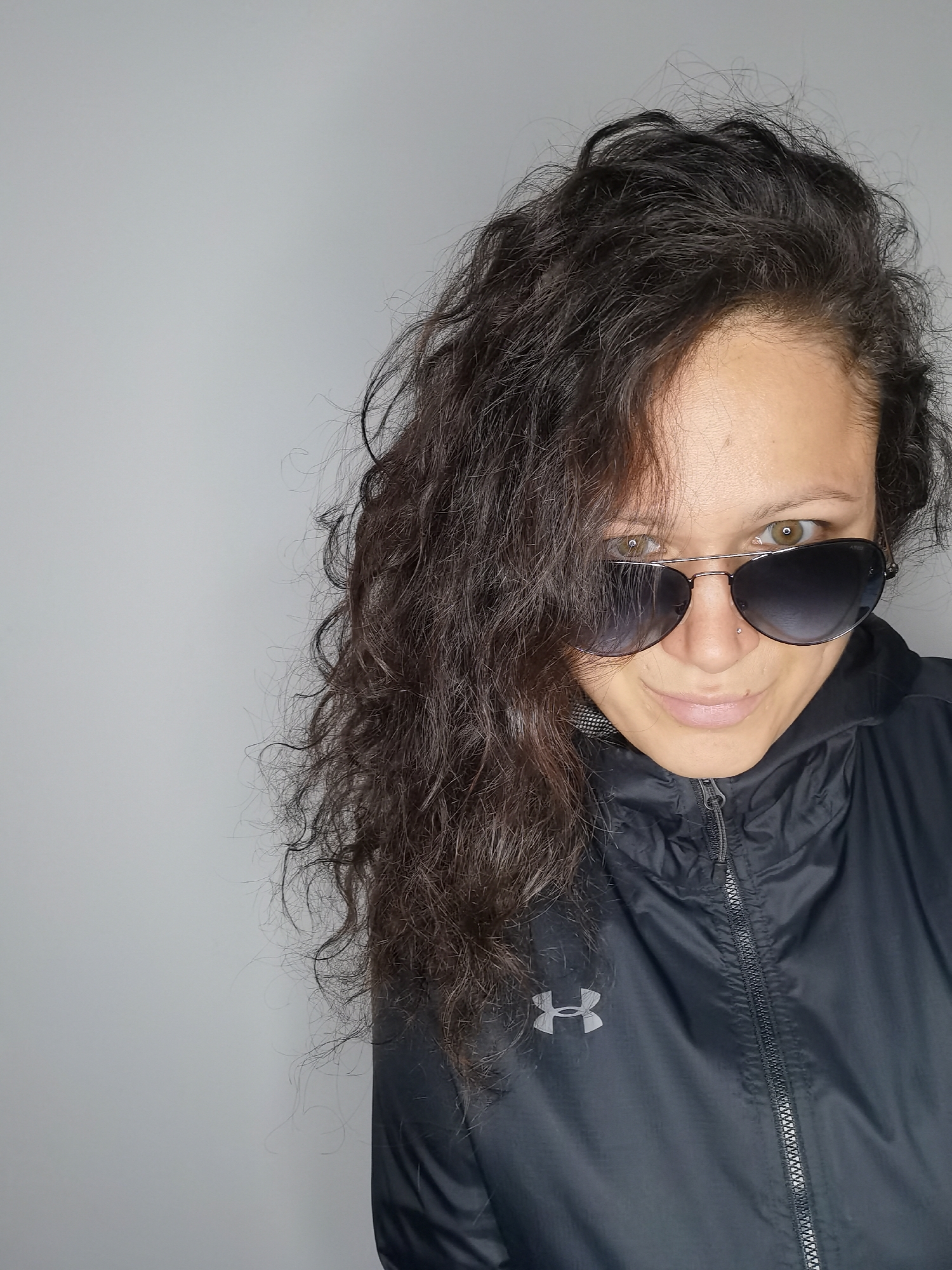 This is a close-up, waist-up, selfie-style photograph of a woman with long, dark, curly hair. She is wearing large, dark aviator-style sunglasses with a thin, silver frame, and a black hooded jacket with a zipper. The jacket has the Under Armour logo on the left breast. Her hair falls over her face, partially obscuring her left eye and cheek. Her gaze is directed at the camera, and she has a slight smile. The background is a plain, light gray wall. There are no clear indicators of the time of day, weather, or specific location beyond the provided context of Chaikovsky, Russia. The overall mood appears casual and confident.