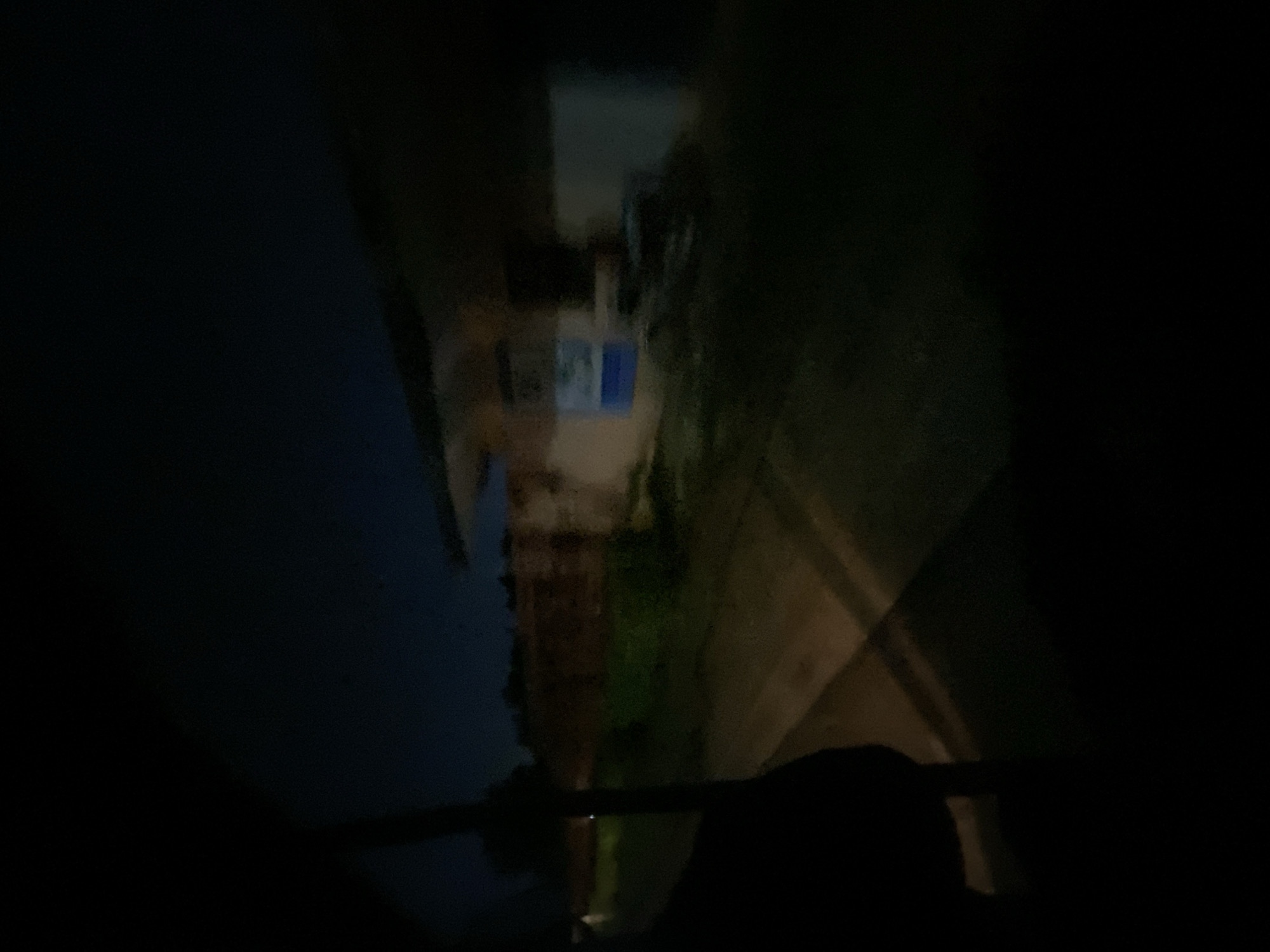 A dark, blurry exterior scene is captured at night, suggesting a view from inside a vehicle. In the background, a building with a covered overhang and an orange-brown brick wall is visible. A portion of a light blue sign with white lettering is affixed to the building's facade. The foreground is dominated by darkness, with a diagonal shadow cast across a paved surface. The overall impression is one of low visibility, characteristic of nighttime photography in an urban or semi-urban setting. The location within Gura Top, Nigeria, is not discernible from the visual information provided.