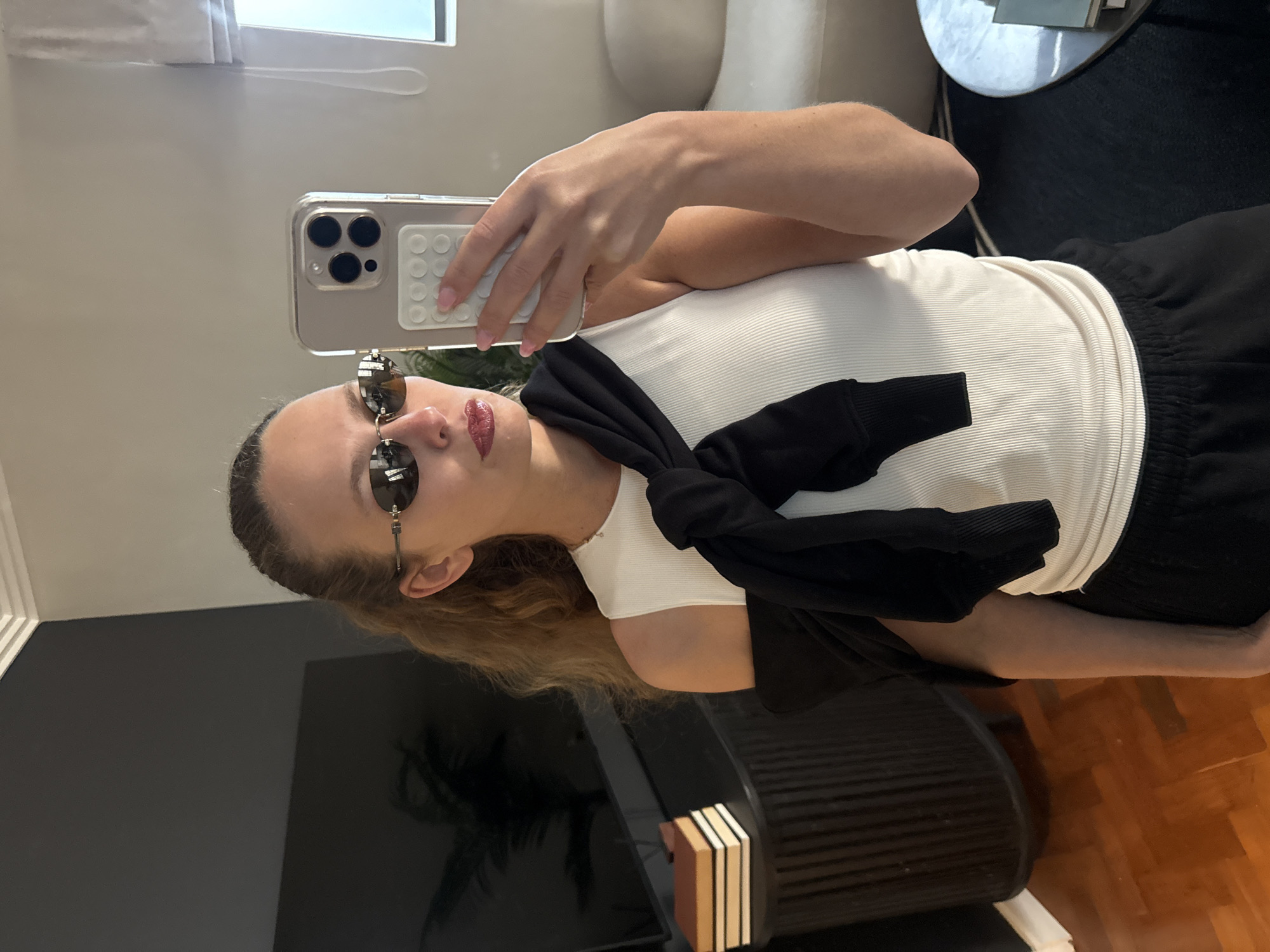 The original image is rotated 90 degrees counter-clockwise. When viewed upright, it depicts a young woman taking a mirror selfie in an indoor setting, likely a residence in Cape Town, South Africa. The woman, with long light brown hair pulled back, wears dark, round sunglasses and has glossy, reddish lipstick. She is dressed in a white ribbed tank top and has a black sweater or cardigan tied around her waist. Her lower body is clad in dark-colored, possibly black, casual pants or shorts.
She holds a light-colored smartphone, likely an iPhone given its triple camera system, in her right hand. The phone is encased in a clear cover, which has a white, rectangular pop-socket or grip accessory with a grid pattern attached to its back. She maintains a neutral expression while posing for the photograph.
The mirror's reflection reveals parts of the room. Natural light streams through a window with white blinds visible in the upper background. To the right, white upholstered furniture and a round table with a dark surface are reflected. In the lower part of the scene, a large, dark, reflective screen, possibly a television, is visible. Adjacent to it, a dark piece of furniture holds a stack of small, rectangular items, resembling books or decorative boxes, in shades of white, beige, and brown. The flooring to the right is warm-toned wooden parquet. There is no visible text in the image.