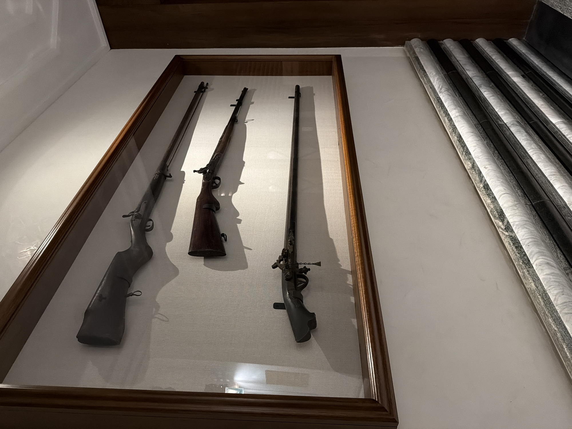 The image shows a display case containing three antique firearms mounted on a light-colored fabric backing, likely in a museum or historical exhibit. The firearms are various types of muskets or rifles, suggesting a collection of historical weaponry. The display case has a dark wood frame. Adjacent to the display case, there are several decorative marble or stone columns, adding to the museum-like setting.