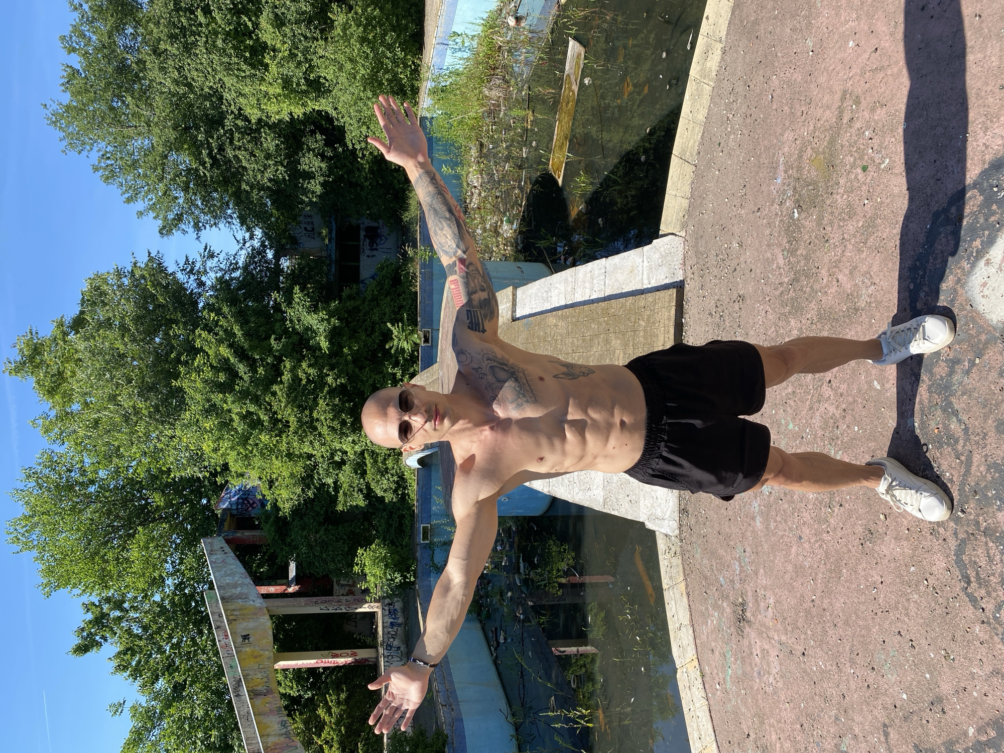 A shirtless, bald man with sunglasses and extensive arm tattoos stands with his arms outstretched in front of an abandoned, overgrown swimming pool. The pool is filled with murky water, debris, and aquatic plants. Behind the pool, a concrete structure covered in graffiti rises amidst dense green trees. The man is wearing black shorts and white sneakers. The ground around him is a cracked, reddish-pink surface with scattered debris and shadows cast by the trees. The scene appears to be in an outdoor, urban-decay setting, possibly the abandoned pool of the Pleven Park Hotel in Pleven, Bulgaria, given its characteristic architecture and state of disrepair.