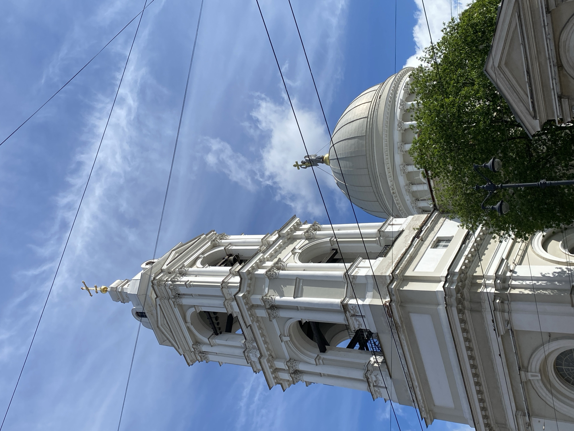 The image shows the architectural elements of the Saint Petersburg’s Saints Peter and Paul Cathedral, Russia, against a backdrop of a blue sky with white cirrus clouds. Power lines are visible across the sky. The cathedral’s bell tower is prominently featured, ascending to a golden cross at its peak. A dome with a statue is also partially visible. Trees are visible on the side of the building.