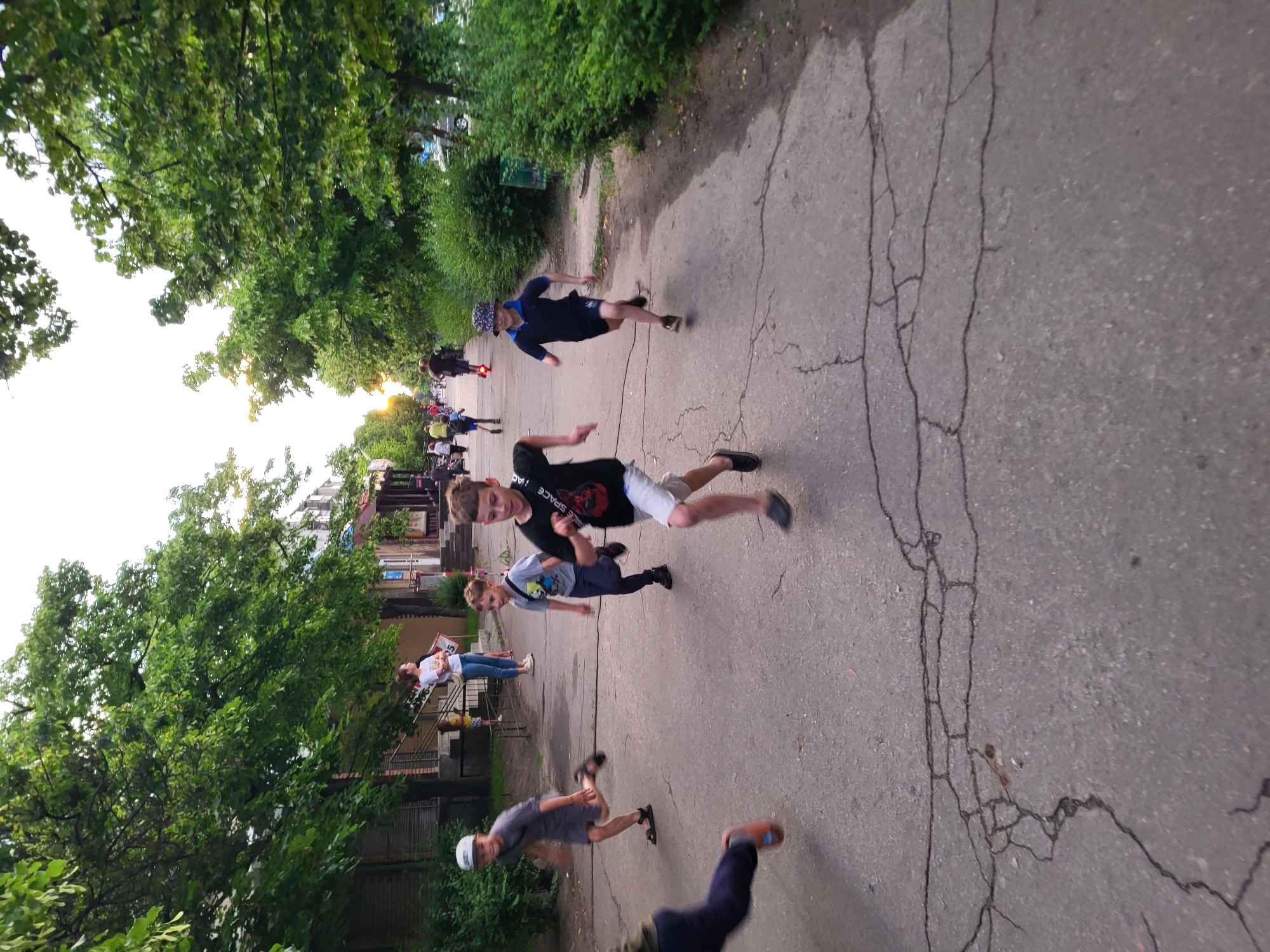 The image depicts a group of children running on an asphalt path in a residential area of Horishni Plavni, Ukraine. Several children are in the foreground, actively engaged in running. One child in the center foreground is wearing a black t-shirt with a graphic and white shorts. To his left, another child in a grey t-shirt and dark pants is also running. Further to the left, a child in a grey t-shirt and sandals is running. On the right side of the frame, a child in a dark blue polo shirt and shorts with a patterned hat is running. In the background, more children and adults are visible. The environment includes trees with green foliage lining the path, and buildings with storefronts and entrances are visible on the left side. The asphalt surface is cracked and weathered. The lighting suggests it is late afternoon or early evening, with the sun setting in the distance.
