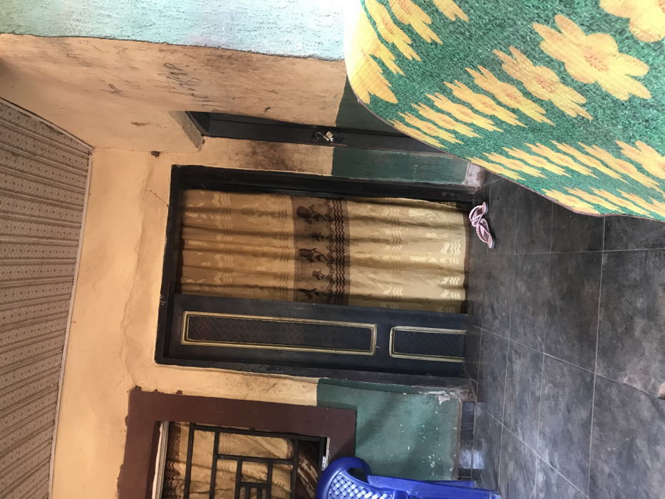 A residential entry presents a dark door with a curtain. A smaller, grated window sits below it. To the right, dark gray tiled flooring with a pair of pink flip-flops is partially covered by a green and yellow floral mat. A blue plastic chair is visible near the bottom. Walls are painted in varying colors including beige, green and blue. The ceiling appears unfinished.