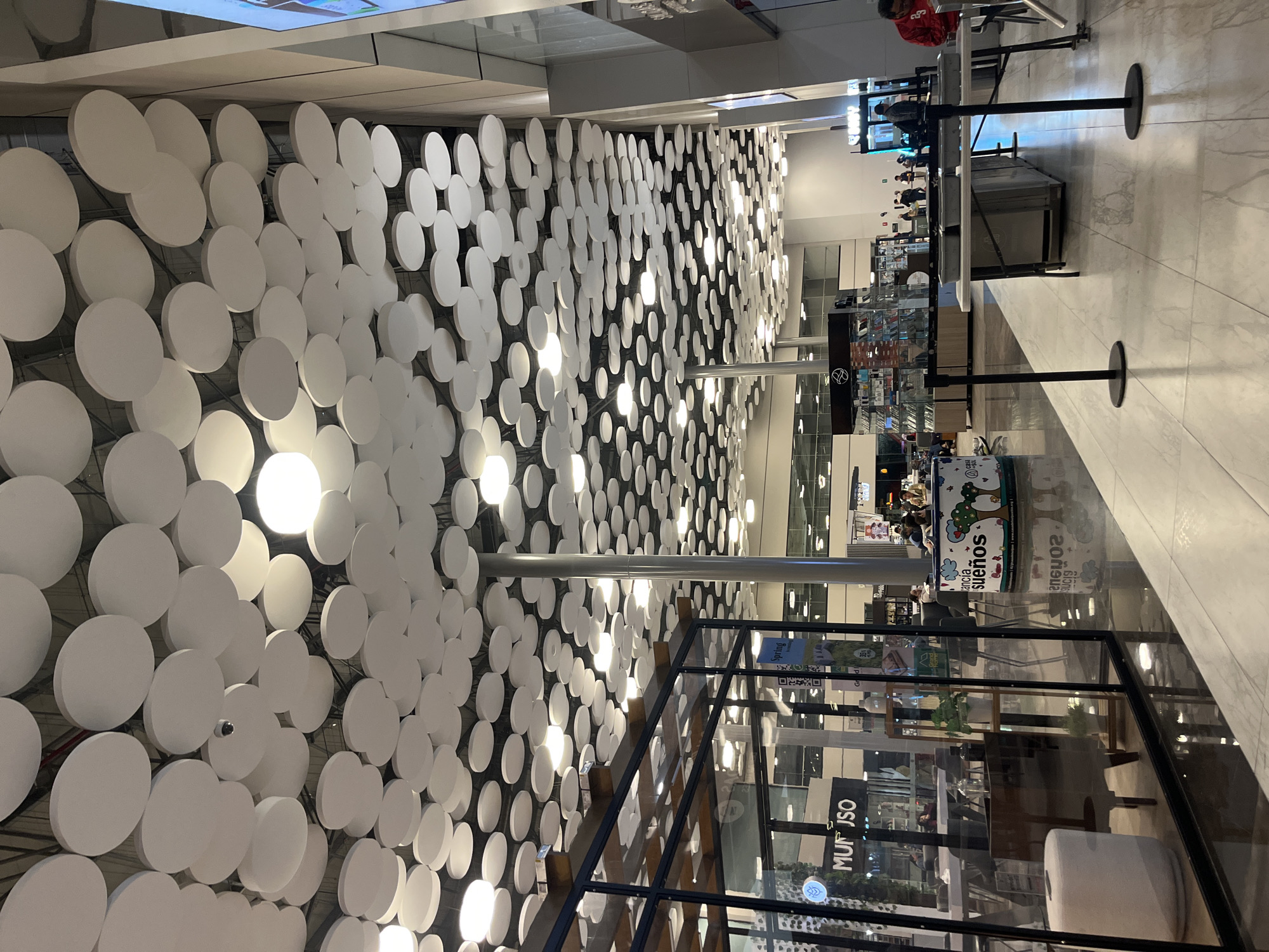 The image displays the interior of a large, brightly lit shopping mall located in Puebla, Mexico. The most prominent architectural feature is the ceiling, which is extensively covered with numerous overlapping, white, circular acoustic panels or decorative discs. Many of these discs integrate recessed light fixtures, providing ambient illumination, and a surveillance camera is visible among them.
The floor consists of highly polished, reflective light-colored tiles. Several tall, circular, metallic-gray support columns extend from the floor to the ceiling.
On the left side of the frame, a glass-fronted retail store with dark framing is visible. The store's interior displays furnishings including a white cylindrical stool, a wooden shelving unit holding a potted plant, and dark seating. A "MINISO" sign is discernible inside the store.
In the midground, centered around one of the columns, stands a large, cylindrical kiosk or display unit. This unit is decorated with cartoon-like illustrations of trees and houses and features text identifying it as "Estancia sueños" and "Casa del Aguacate Puebla."
On the right side of the frame, a white folding table is set up with three rectangular metal trays on its surface. Two black stanchions with round bases are positioned on the floor in front of this table. A person wearing a red long-sleeved shirt is partially visible on the far right, near what appears to be an entrance or gate.
Several individuals are present throughout the mall space, primarily in the midground and background, engaged in walking or standing, consistent with general public activity within a commercial environment.