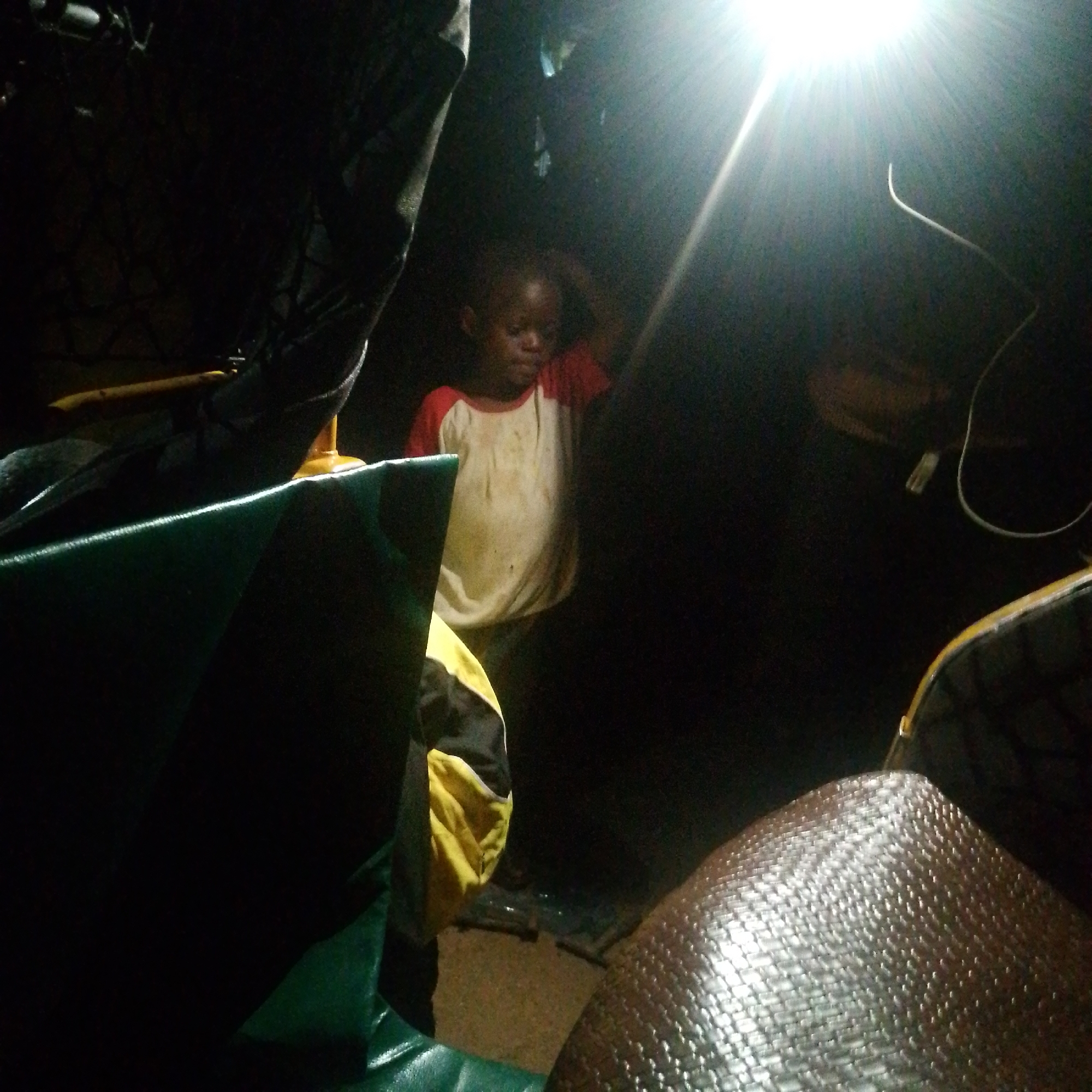 This image depicts a dimly lit, enclosed space, likely at night, illuminated primarily by a single, intensely bright artificial light source positioned in the upper right. The light casts strong rays and deep shadows across the scene, creating a stark contrast. A white wire or cord is visible near the light source.
The central subject is a young, dark-skinned child, likely a boy, standing in the middle distance. He wears a white t-shirt with red sleeves that appears somewhat soiled or stained. One of his hands is raised to the side of his head, possibly adjusting his hair or scratching. His face is partially obscured by shadows and the bright light, and his expression appears neutral.
In the foreground, to the left, there is a large, dark green, textured surface, possibly a tarp or a seat cover. Next to it, a folded piece of yellow and black fabric or clothing is visible. To the right, another textured surface, possibly woven or corrugated in a metallic brown/silver hue, forms part of the immediate foreground. Other indistinct shapes and materials, including a yellow bar-like object, are visible within the deep shadows.
The overall setting appears to be a humble or temporary environment, possibly a small room, a tent, or the interior of a vehicle, given the raw materials and the reliance on a single artificial light source. No specific activity is taking place beyond the child standing. No emotions are clearly discernible, and there is no visible text. The location is unknown.