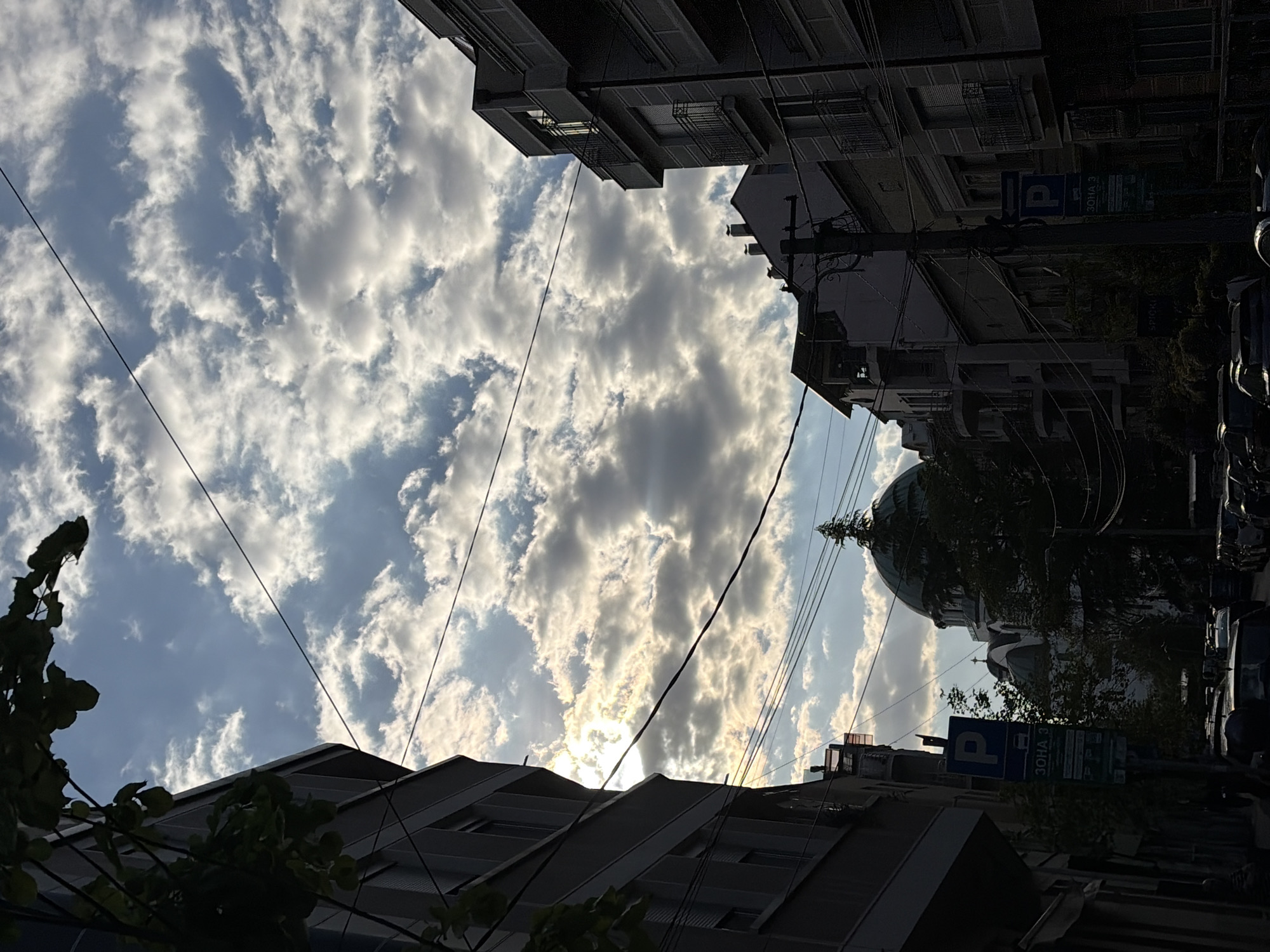 The image presents an upward perspective of an urban environment, capturing a dramatic sky framed by buildings. The scene depicts a street in Belgrade, Serbia.
The central and upper portions of the frame are dominated by a dynamic sky, filled with large, bright white and gray cumulus clouds. Sunlight, largely obscured by the clouds, breaks through in brilliant rays, creating a striking "god rays" effect towards the lower middle of the sky.
On the left and bottom left, residential or commercial buildings rise, their facades and multiple balconies visible in partial silhouette against the bright sky. Dark green foliage from trees or bushes partially obscures the lower parts of these structures.
On the right side of the frame, more buildings are present, mostly in shadow. A distinctive large, dark domed structure, likely a church or cathedral, with a small spire at its apex, stands prominently on this side. A network of dark utility wires and power lines crisscrosses the sky throughout the image, adding to the urban feel.
A blue parking sign is clearly visible on the right, displaying the white letter "P" and the Cyrillic text "ЗОНА 3" (ZONA 3), indicating a designated parking zone. Some parked cars are faintly discernible beneath the buildings on the right, largely engulfed in shadow.
The lighting suggests late afternoon or early evening, characterized by the low sun angle and the interplay of intense light and deep shadows. The overall impression is one of a serene yet dynamic urban landscape, with the majestic cloudscape as the focal point.