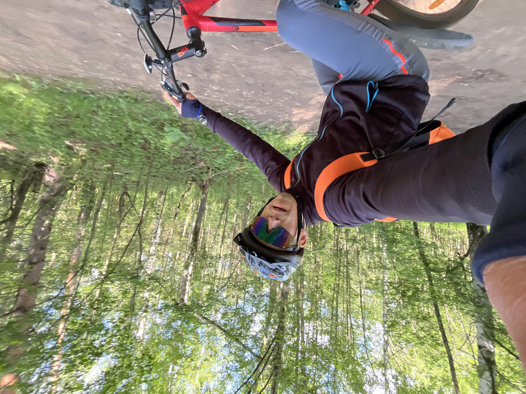 This first-person point-of-view image captures a male cyclist on a sunny day in a forest, likely in Chaikovsky, Russia. The cyclist, seen from the chest up, is wearing a dark long-sleeved cycling jersey with bright orange and light blue accents, grey cycling pants with orange zippers, and a dark helmet with a visor. He sports distinct rainbow-colored reflective sunglasses. His right hand is on the handlebar of a red mountain bike, part of which is visible, including the front wheel and handlebar controls. Orange straps are visible across his chest and shoulder, suggesting he is wearing a backpack or hydration vest.
The background features a lush green forest with numerous tall trees and dense foliage, indicating a vibrant, healthy ecosystem. A dirt or gravel path is visible beneath the bicycle. Sunlight filters through the tree canopy, creating bright patches and suggesting a clear sky. The overall impression is of an enjoyable outdoor recreational activity like mountain biking on a bright, pleasant day. A small, indistinct red logo or text, possibly "GS," is barely visible on the bicycle's frame near the handlebar stem.