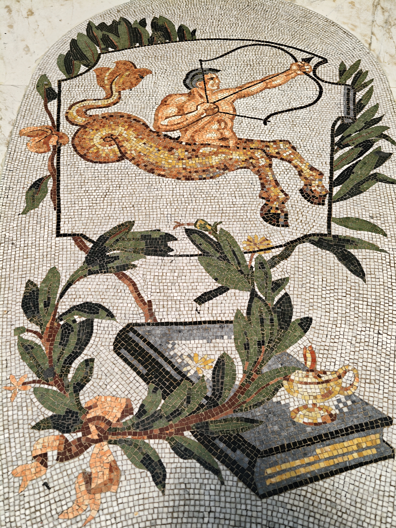 This is a detailed mosaic image depicting a centaur archer drawing a bow and arrow. The centaur has a human torso with muscular arms and a human-like face, and the body of a horse. It is positioned in a leaping or galloping pose, with its rear legs extended and front legs bent. The centaur is looking towards the right of the frame. In the foreground, there are floral elements, including green leaves, brown branches, and orange flowers. There is also a depiction of an open book with a mosaic lamp resting on it, and a cylindrical object that could be interpreted as a scroll or another book. The mosaic is set against a white tiled background.
This mosaic is located on the exterior of the Museo Archeologico Nazionale di Napoli (National Archaeological Museum of Naples) in Naples, Italy. Specifically, it is part of the architectural decoration of the museum's facade.