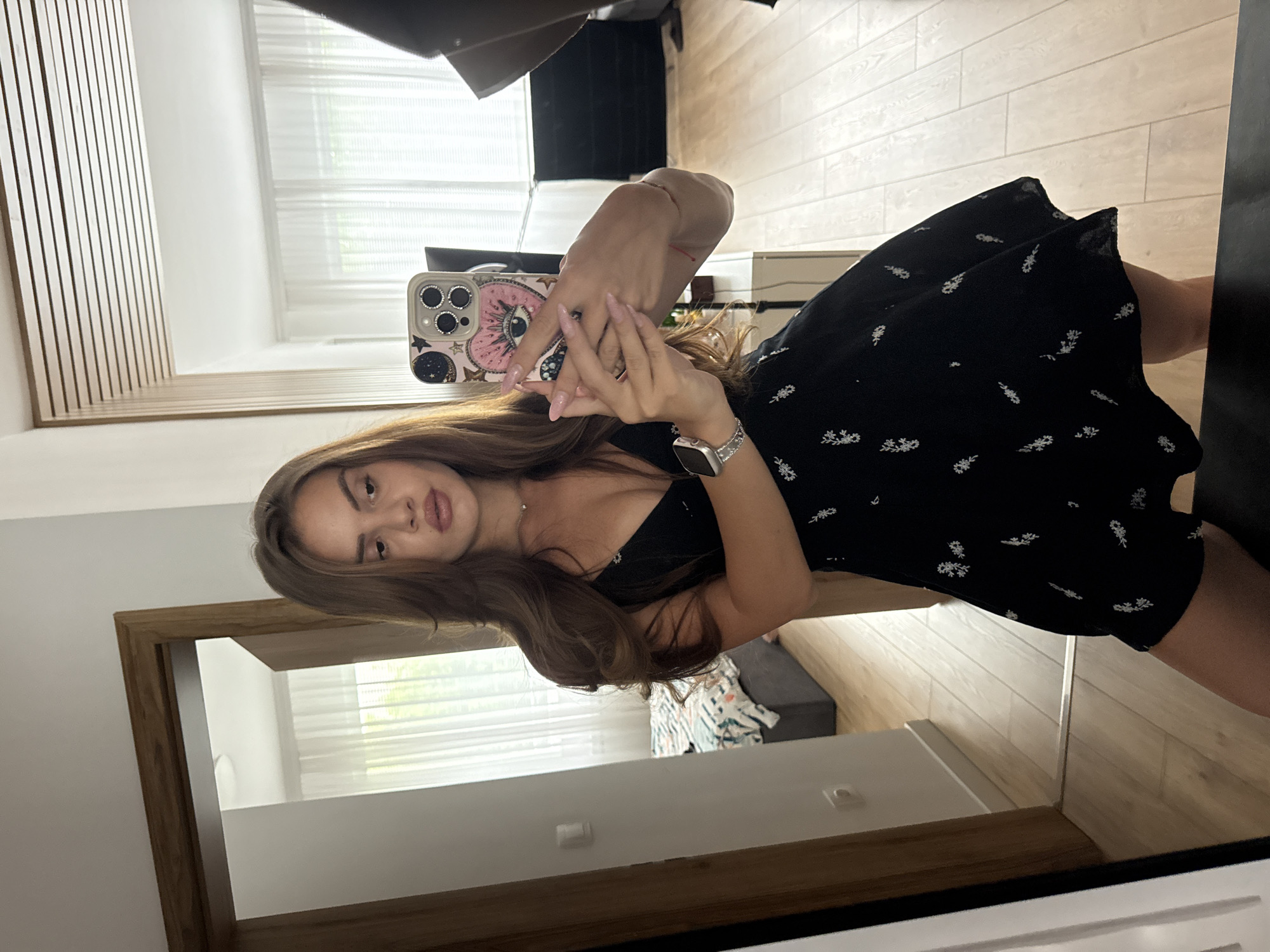 A woman with long, brown hair takes a mirror selfie in what appears to be a bedroom. She is wearing a short, black dress with a floral pattern and a v-neckline. She is holding a smartphone with a decorative case in her right hand, which is raised to take the photo. Her left arm is bent at the elbow. She is wearing a watch on her left wrist. The background includes a mirror, a wooden door frame, a bed with patterned bedding, and white curtains covering a window. The floor is made of light-colored wood planks. The lighting suggests an indoor setting, possibly during the daytime.