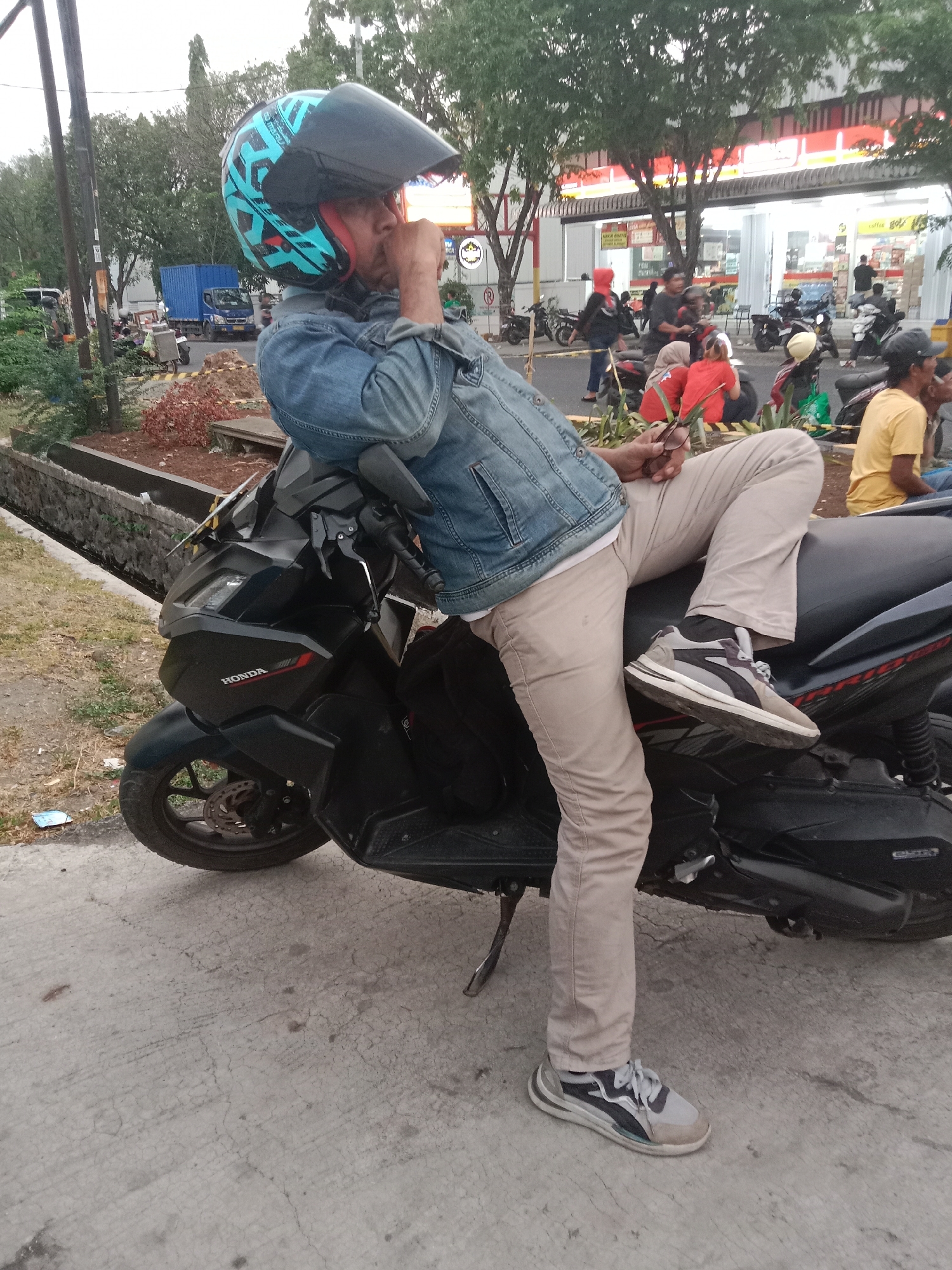 A person is sitting on a black scooter, wearing a blue denim jacket, beige pants, and a teal and black helmet. They are holding a pair of glasses in their right hand, with their thumb resting on their chin. The scooter has "HONDA" written on the side. In the background, there are trees, a blue truck, other parked motorcycles, and a building with red and white signage that appears to be a convenience store. The ground is paved concrete, and there is a drain along the left side of the image. The scene suggests an urban environment, possibly in Semarang, Indonesia, based on the presence of the scooter and the architectural style of the building.