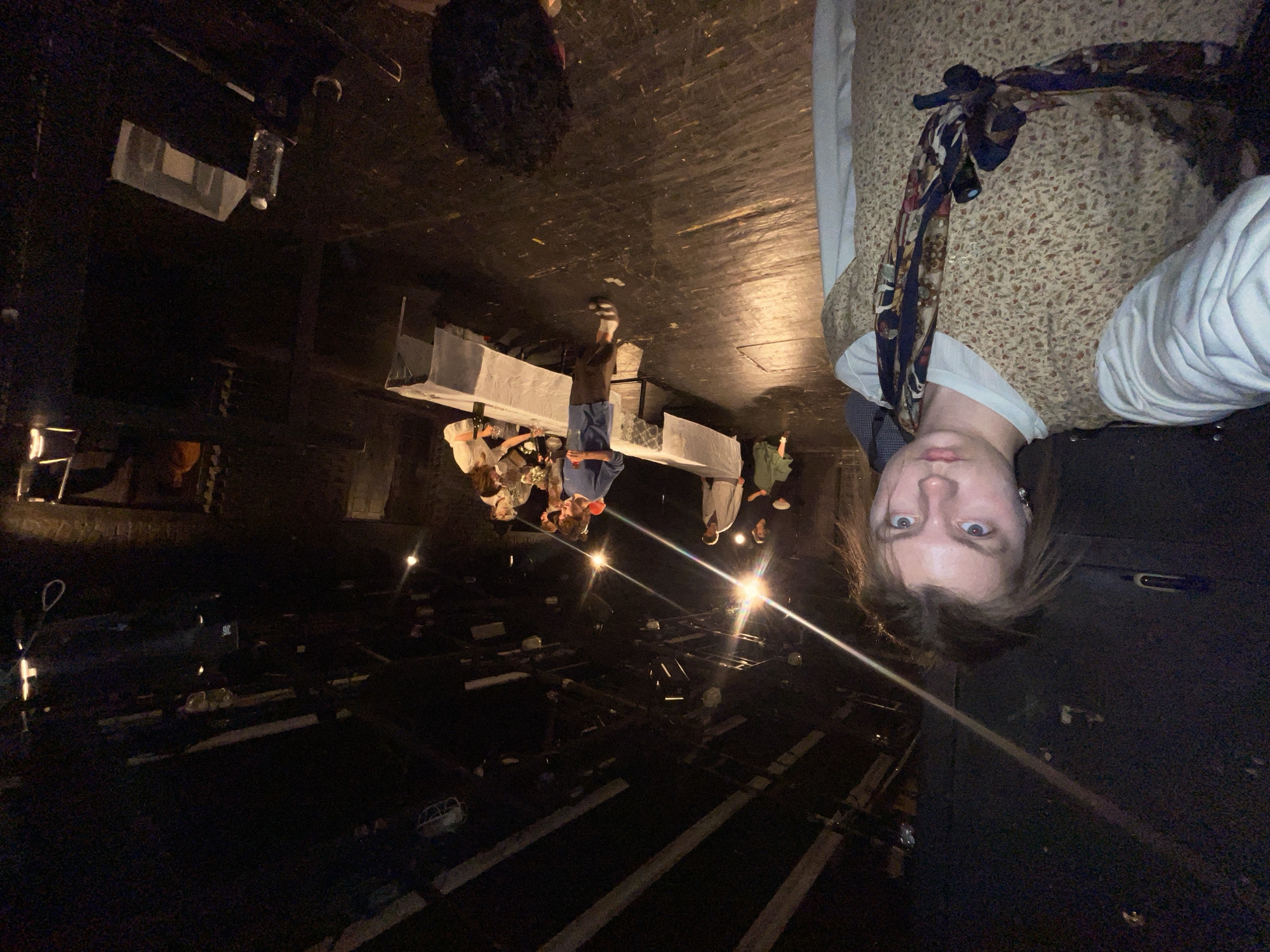 The image shows the interior of what appears to be a theater during a production. The scene is dimly lit, with visible stage lights and dark shadows. There is a group of people around a table set with props, possibly actors in costume rehearsing or performing. A person is taking a selfie in the foreground, wearing a vest and a patterned necktie. The environment suggests a backstage or offstage area due to the exposed beams, structures, and general darkness. There is no evidence to suggest that the picture was taken in Moscow, Russia.