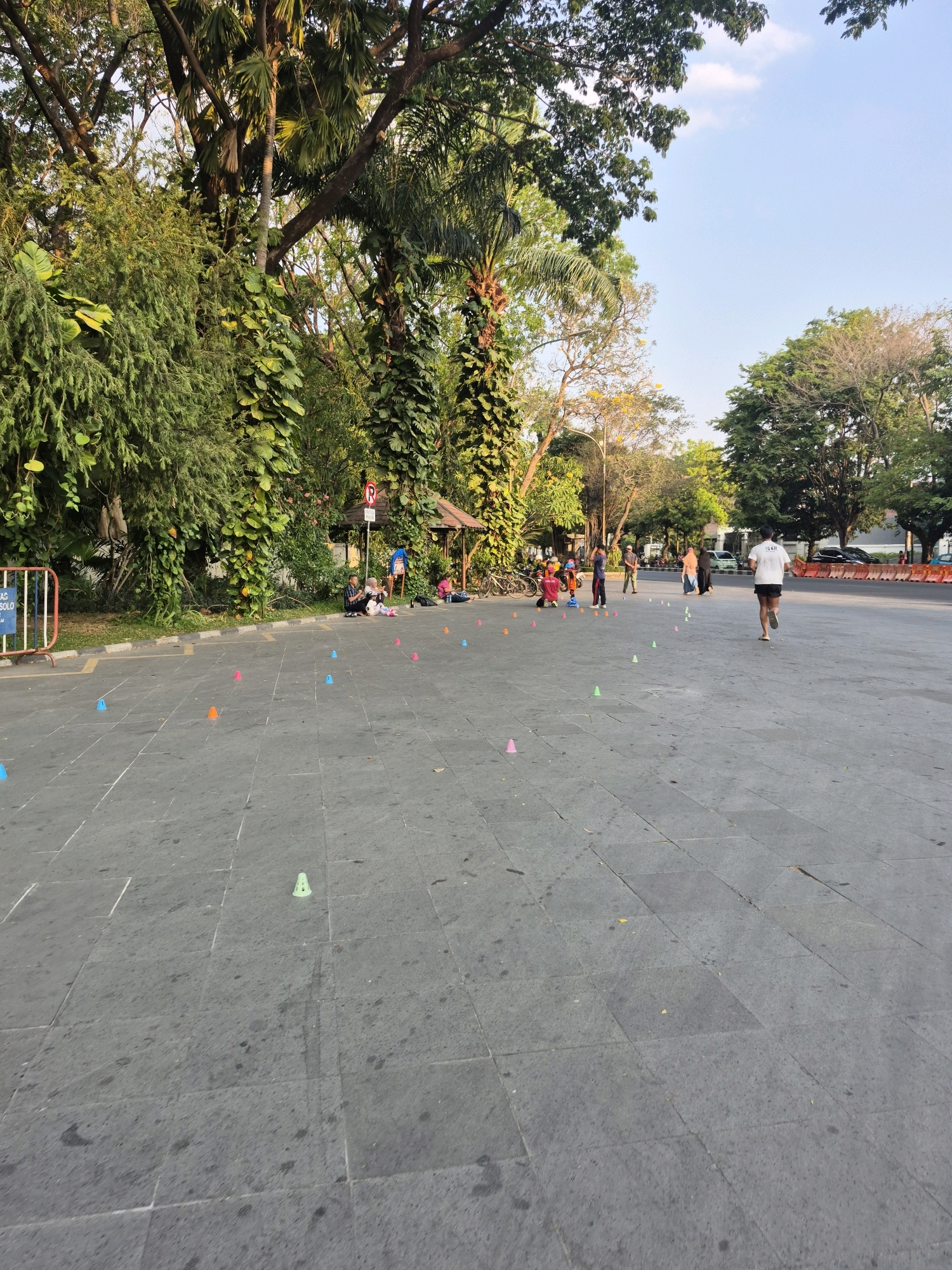 The image depicts an outdoor scene in the city of Surakarta, Indonesia, likely in a public park or recreational area. The foreground is dominated by a paved, grey, stone-tiled surface, possibly a promenade or plaza. Scattered across this surface are numerous colorful traffic cones in various shades of orange, blue, and green, suggesting a training or obstacle course area.
In the mid-ground, several individuals are present. To the left, a group of people are seated on the ground near a lush green hedge and trees. Among them, a person appears to be looking at a phone. Further to the right, a man in a white t-shirt and black shorts is jogging away from the camera. Other individuals, including children, are also visible, some appearing to be engaged in activities near the cones. A small, open-sided shelter with a brown roof is situated in the background, surrounded by dense foliage and tall trees. The background also shows trees with vibrant green leaves and some taller palm-like trees, indicating a tropical or subtropical climate. A clear blue sky with a few white clouds is visible above the trees.
To the far left, a metal barrier with white and blue markings is present, potentially delineating an area or entrance. Beyond the paved area, a road with vehicles, including cars and what appears to be a motorcycle, can be seen, suggesting the proximity of a street. The overall lighting suggests it is daytime, likely late afternoon or early morning due to the warm tones and the angle of the sun. The scene conveys a sense of casual activity and recreation in a park-like urban setting.