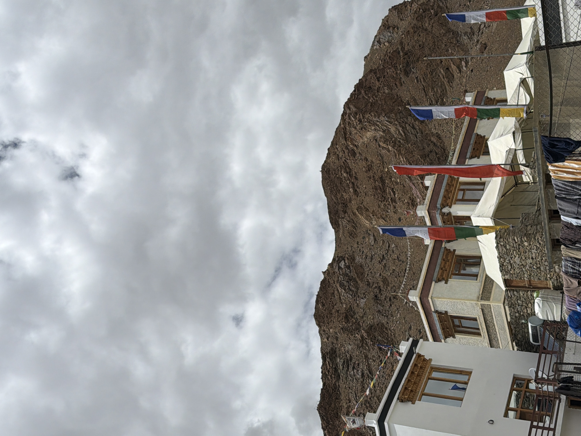 In Karzok, India, under a cloudy sky, a cluster of buildings stands against a rocky mountain. The structures, a mix of white-plastered and stone-faced designs, feature traditional wooden window frames. Prayer flags in various colors flutter between them. In the foreground, tents are set up, and laundry hangs on a line. A satellite dish and water tanks are visible on a lower level, indicating a residential area. The mountain's rugged texture and the muted colors of the scene suggest a high-altitude, arid environment.