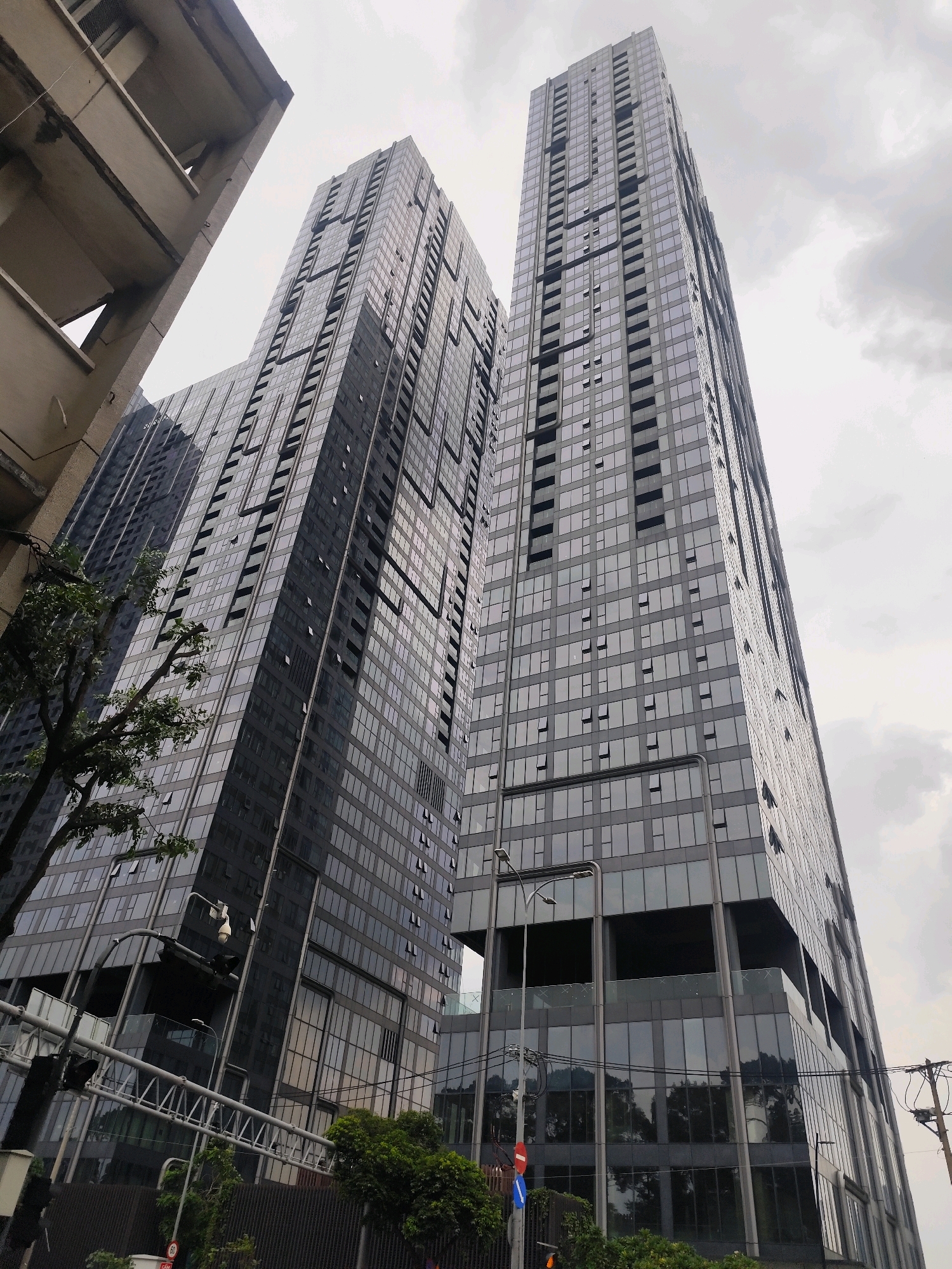 The image shows a view looking up at two modern, tall glass skyscrapers in Ho Chi Minh City, Vietnam. The skyscrapers are adjacent to each other, with one slightly taller than the other. They have a reflective glass facade with many windows, some of which are open. The sky above is overcast and cloudy. In the foreground, a portion of an older, concrete building with balconies is visible on the left, and some trees and street infrastructure, including traffic lights and power lines, are present. The overall scene depicts an urban environment with a contrast between older and newer architecture.