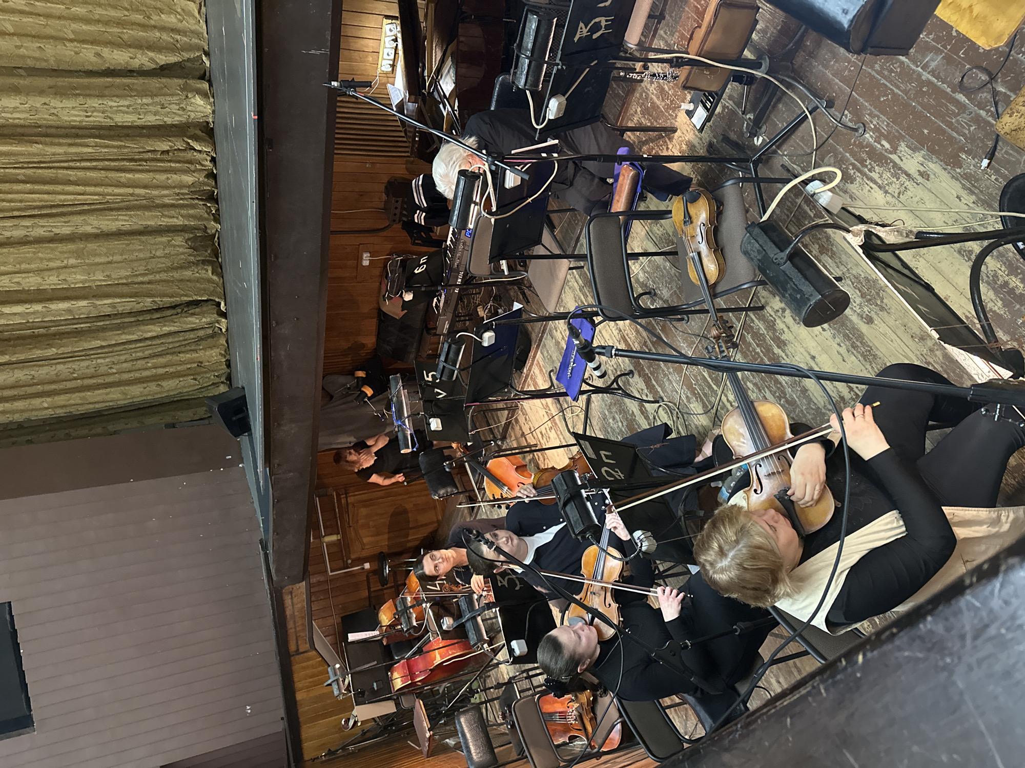 The image depicts an elevated, overhead view of a musical ensemble rehearsal or performance in an indoor venue located in Unknown, Unknown. The scene is dominated by a group of musicians and their instruments arranged on a worn, dark-stained wooden floor, with a stage and curtains forming the background. In the upper portion of the image, a large, deeply pleated, golden-brown curtain hangs across the back wall of a stage. In front of the curtain, a dark, elevated stage platform is visible, with a black speaker placed on its left edge. To the far left, a tall, plain brown wall structure with vertical paneling is present. The main focus is on the musicians. In the left foreground, a woman with light brown hair, wearing a black long-sleeved top and a light beige sleeveless vest, is seated and playing a violin, holding the bow on the strings. Behind her and slightly to the right, another woman with dark hair in a ponytail, wearing a black long-sleeved top, is also seated and playing a violin. Further back and to the right of the first two, a woman with dark hair, wearing a black top with a white collar visible, is seated and playing a cello, with the instrument positioned between her legs. Behind her, a woman with glasses and dark hair, wearing a dark top, is seated and looking towards the right, with a string instrument (likely another cello) partially visible behind her. To the far right of the ensemble, an older individual with white hair, wearing a black long-sleeved top, is seated and playing a black keyboard synthesizer, with sheet music on a stand in front of them. Behind the keyboard player, a dark brown grand piano is partially visible. An oboe or clarinet hangs from a stand to the right of the keyboardist, and a silver flute rests on a blue cloth on a stand further left. Numerous black music stands, many equipped with small lights, are scattered among the musicians. Several microphone stands, including one prominent one in the center foreground, are positioned around the ensemble. The floor is made of distressed, medium-brown wooden planks, showing signs of wear and various darker stains. Electrical cables snake across the floor, connecting instruments and equipment. Black metal chairs provide seating for the musicians. In the background, behind the ensemble, a woman in a black top is standing and observing the group.