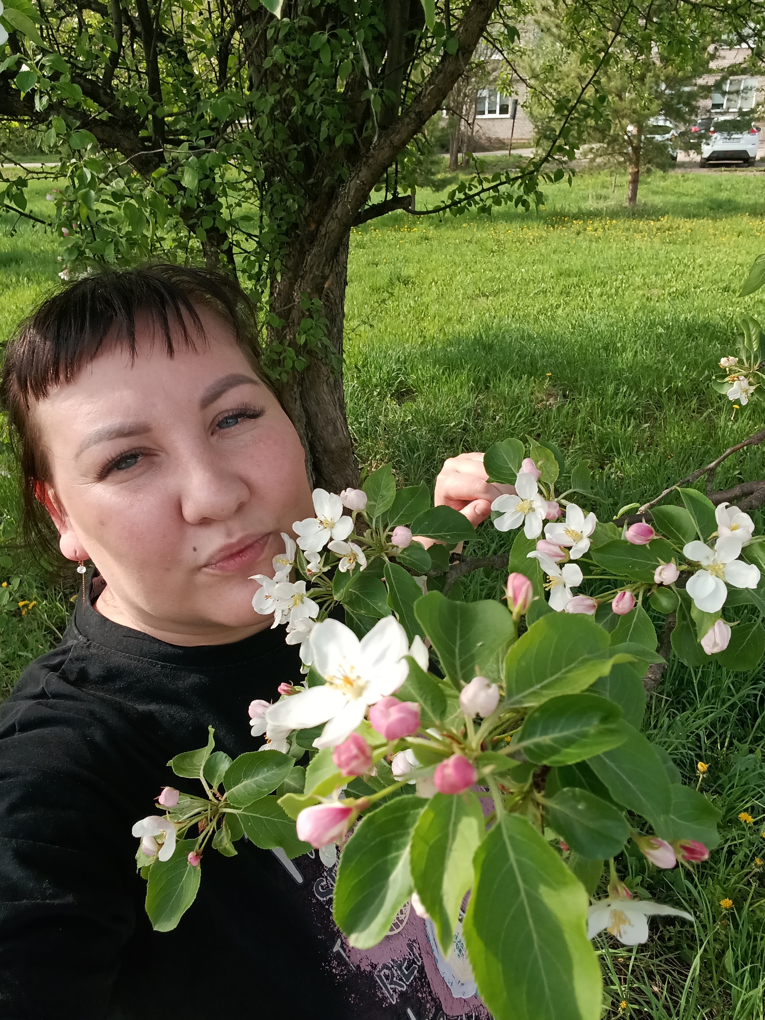 The image is a selfie of a woman with brown hair and bangs, posed next to a blooming tree. She is wearing a black shirt and a small earring is visible on her left ear. She is holding a branch of the flowering tree, with white blossoms and pink buds, close to her face and is making a slight kissing face. The background includes a green grassy area, with a tree trunk behind the woman. In the distance, there are buildings and parked cars. The scene suggests a park-like setting in the city of Osa, Russia, during springtime when trees are in bloom.