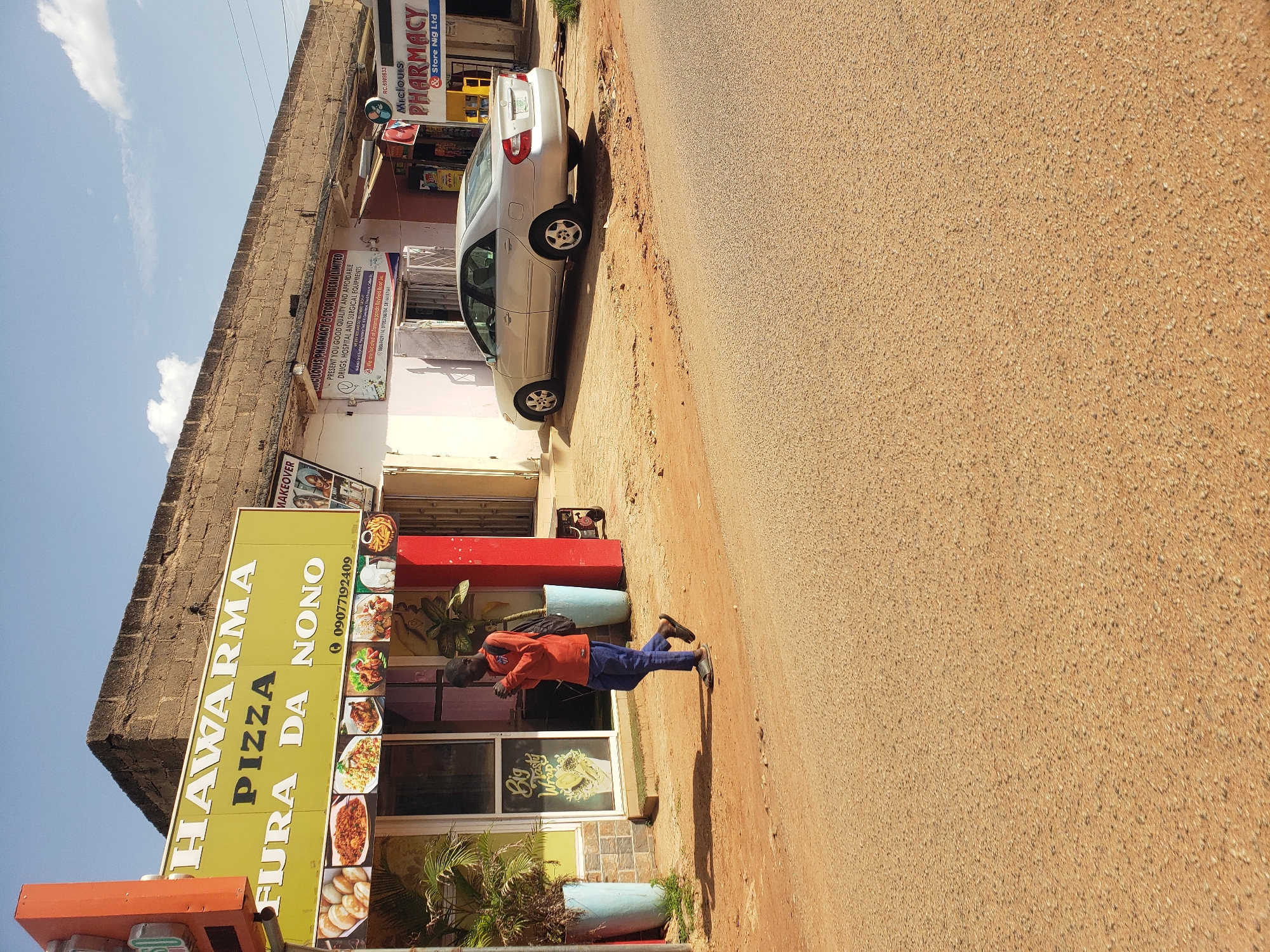 In Gura Top, Nigeria, a street scene unfolds under a bright sky. A person in an orange long-sleeved shirt and blue trousers walks across a dirt road towards a building with a sign advertising "HAWARMA PIZZA FURA DA NONO" and a phone number "09077192409". Images of food are displayed on the building's facade. Adjacent to this, another establishment, "MICLOIS PHARMACY & STORE NIGERIA LIMITED," displays an advertisement for drugs, hospital, and surgical equipment, with a phone number "08064297116" and "07032302364." A silver sedan is parked on the street, casting a shadow. The environment appears dusty with scattered debris.