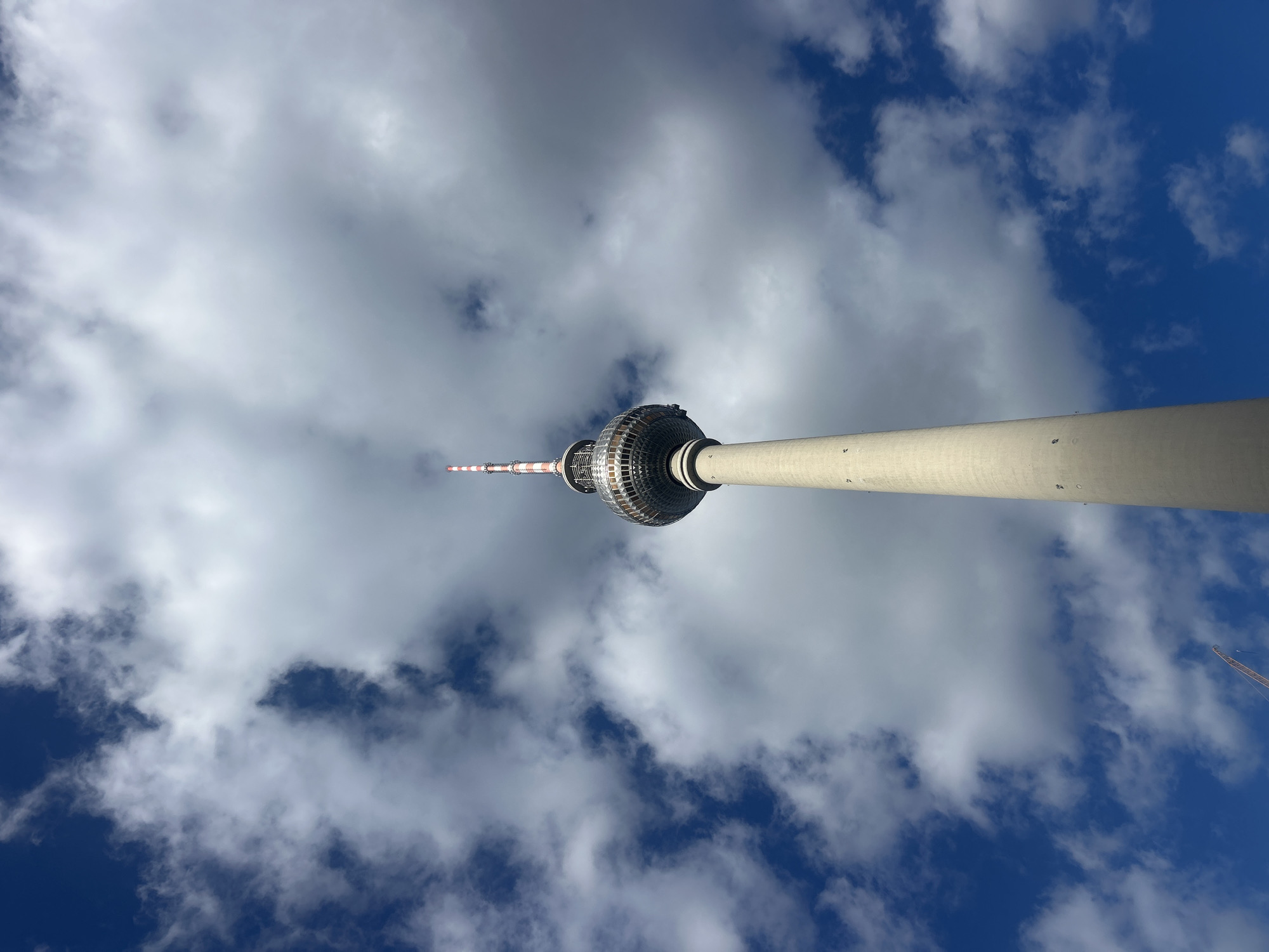 The image shows a low-angle, upward shot of the Fernsehturm (Television Tower) in Berlin, Germany. The tower, a prominent landmark, is a tall, concrete structure with a silver spherical observation deck and a red and white striped antenna at the very top. The tower is set against a dramatic sky filled with a mix of bright blue patches and large, textured white and grey clouds. The clouds are dynamic and somewhat turbulent, creating a sense of depth and movement in the sky. Part of a construction crane is visible in the lower left corner of the image. The lighting suggests it is daytime, with sunlight illuminating the tower and the clouds, casting subtle shadows.